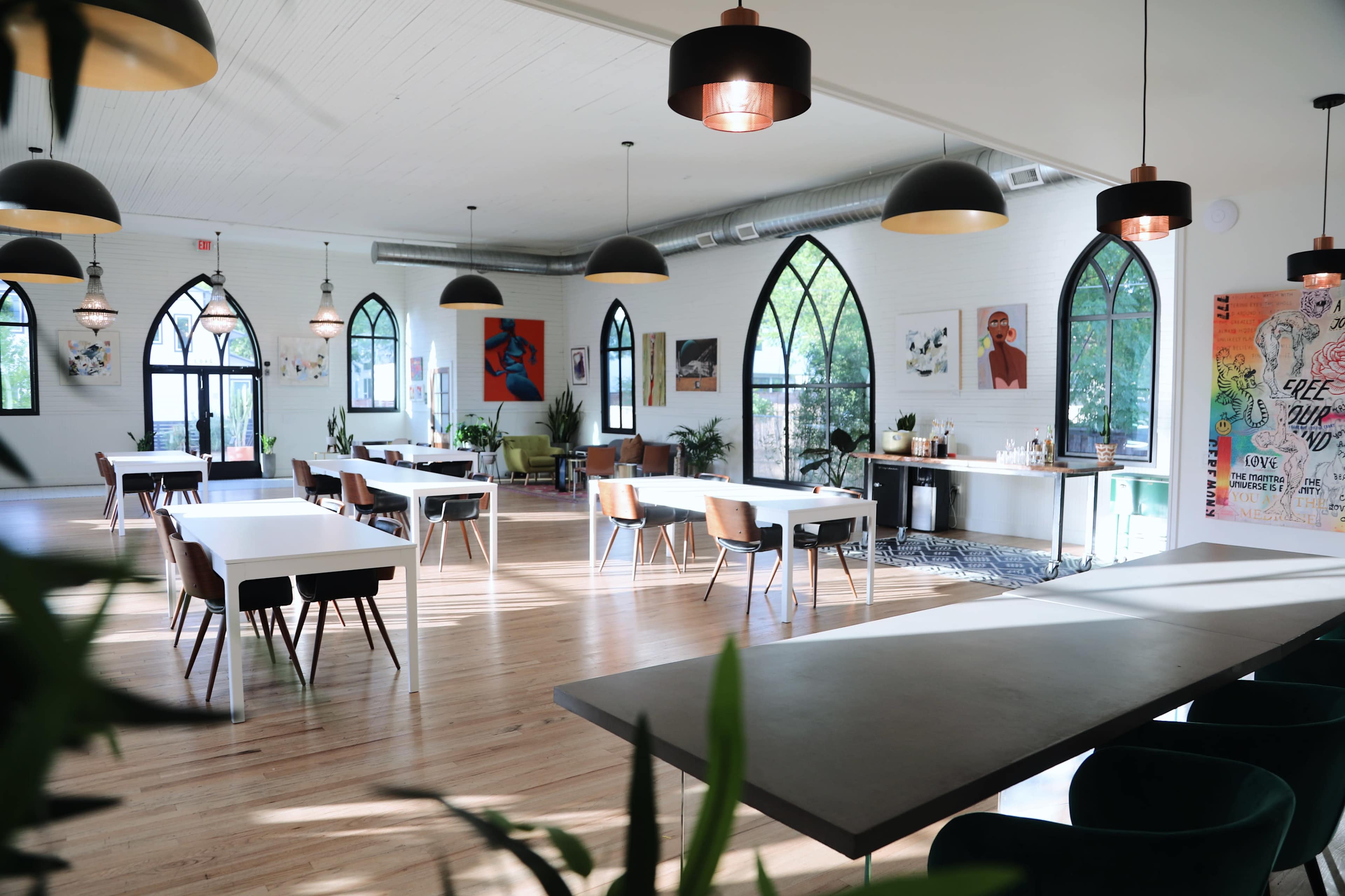 A spacious, well-lit room featuring several tables and chairs, large windows with arched designs, and various artworks on the walls.