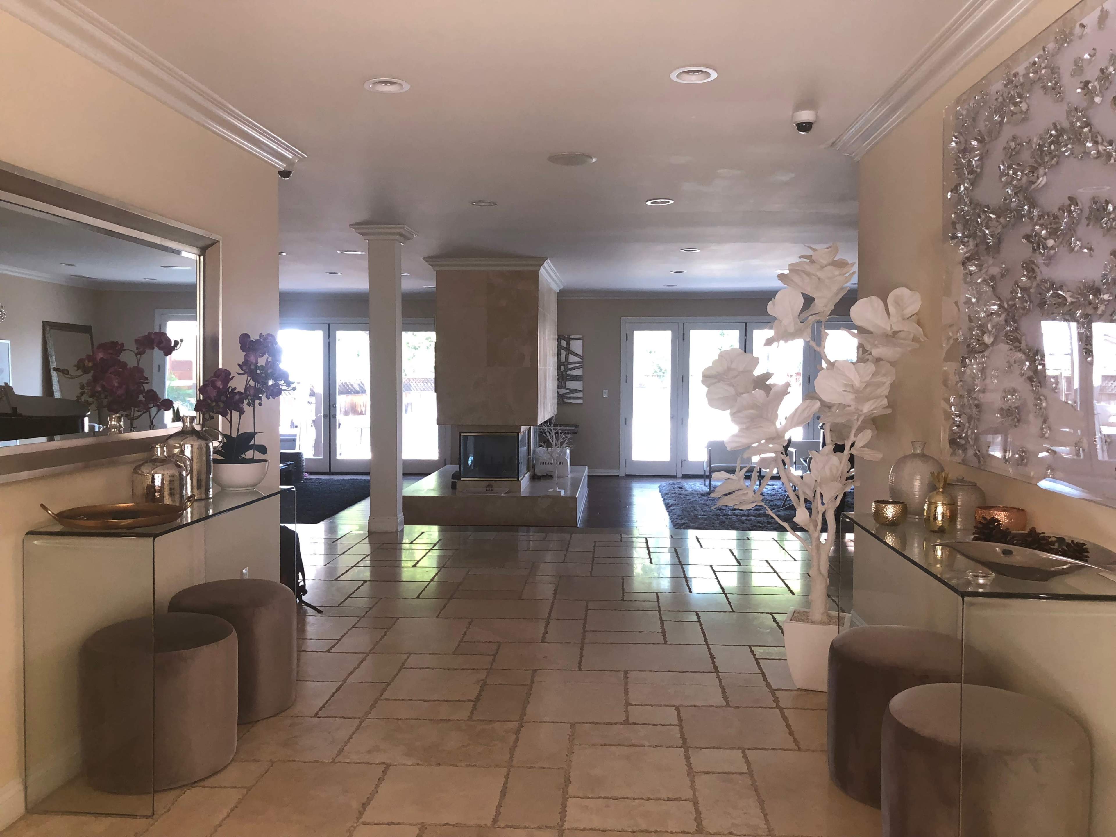 The image shows a spacious, well-lit hallway with tile flooring, a central fireplace, glass tables, and decorative elements including white plants and flower arrangements.
