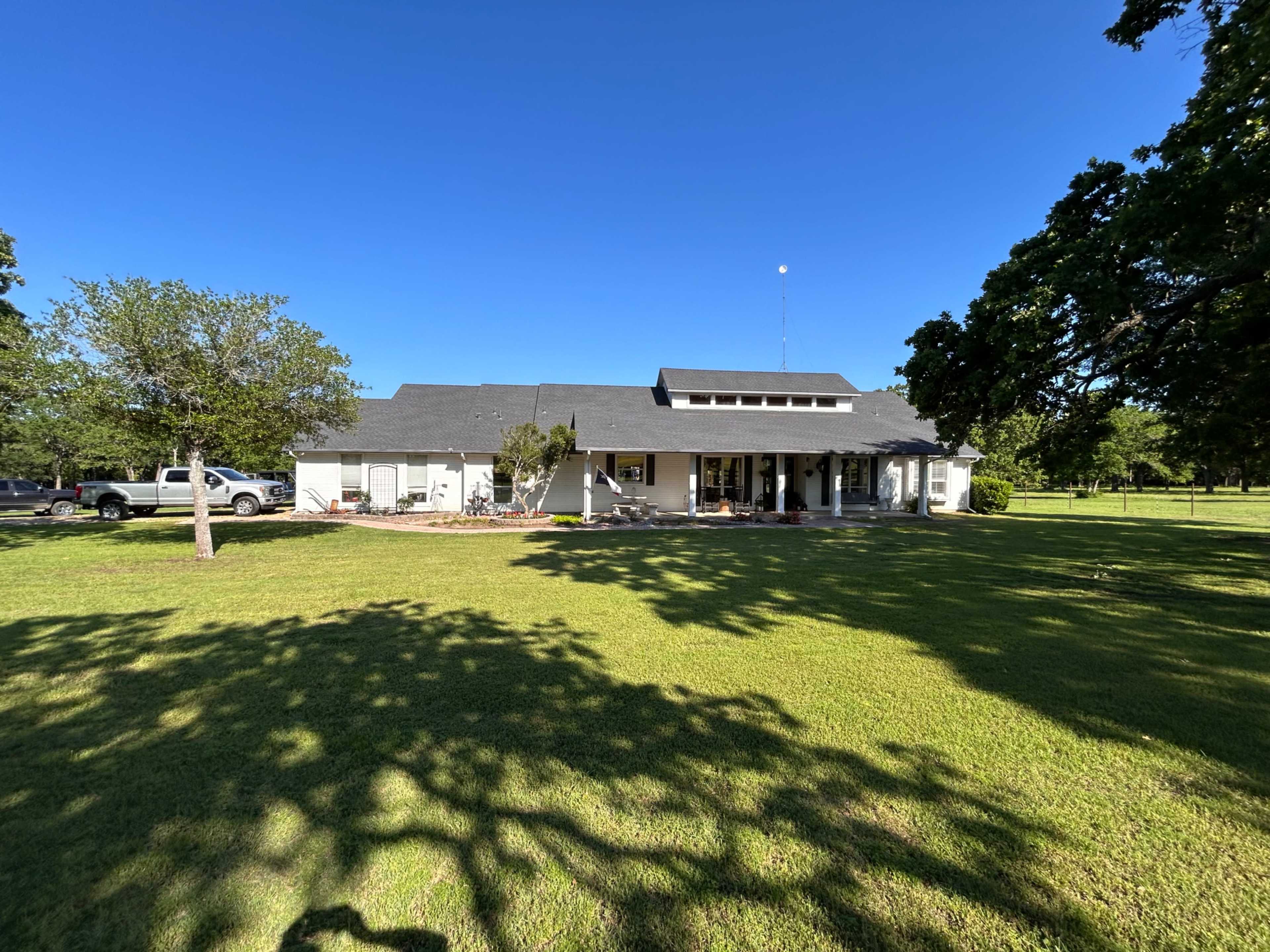 A single-story house with a grey roof and front porch is set on a green lawn surrounded by trees and a gravel driveway.