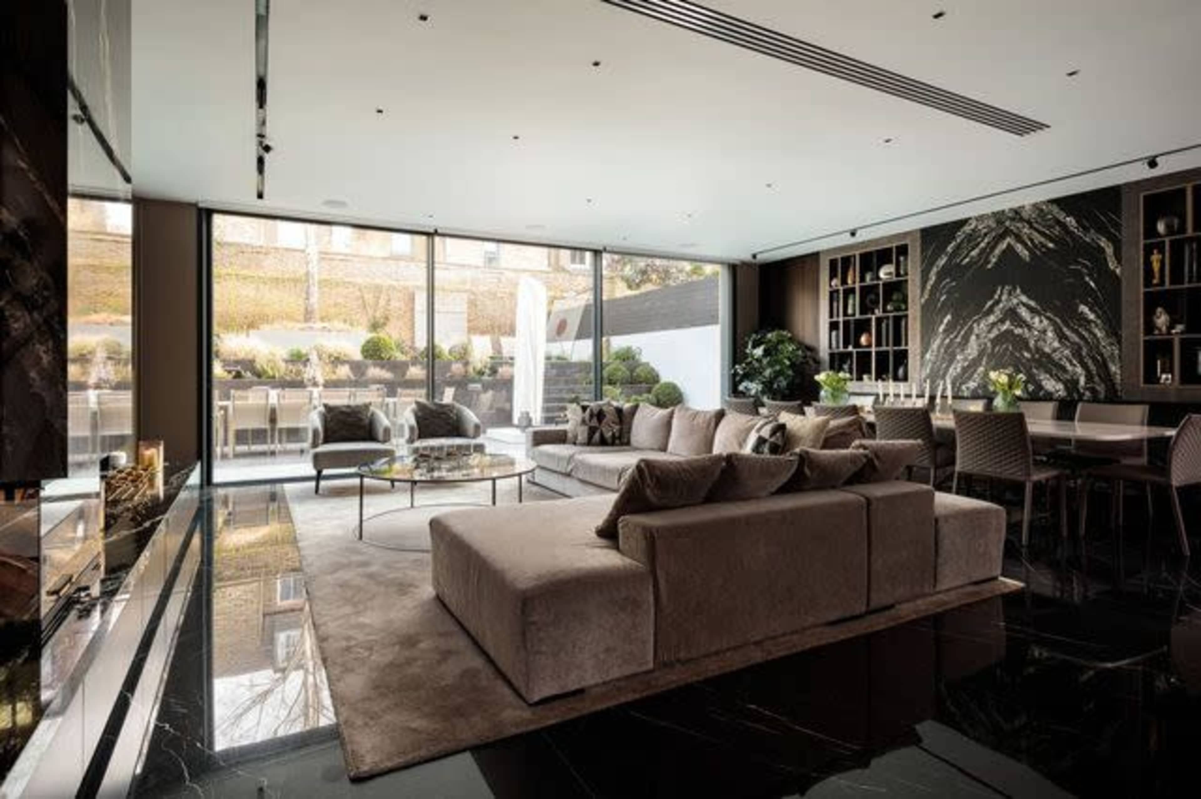 The scene features a modern living room with a large sectional sofa, a glass coffee table, and an open view of an outdoor patio through floor-to-ceiling windows.