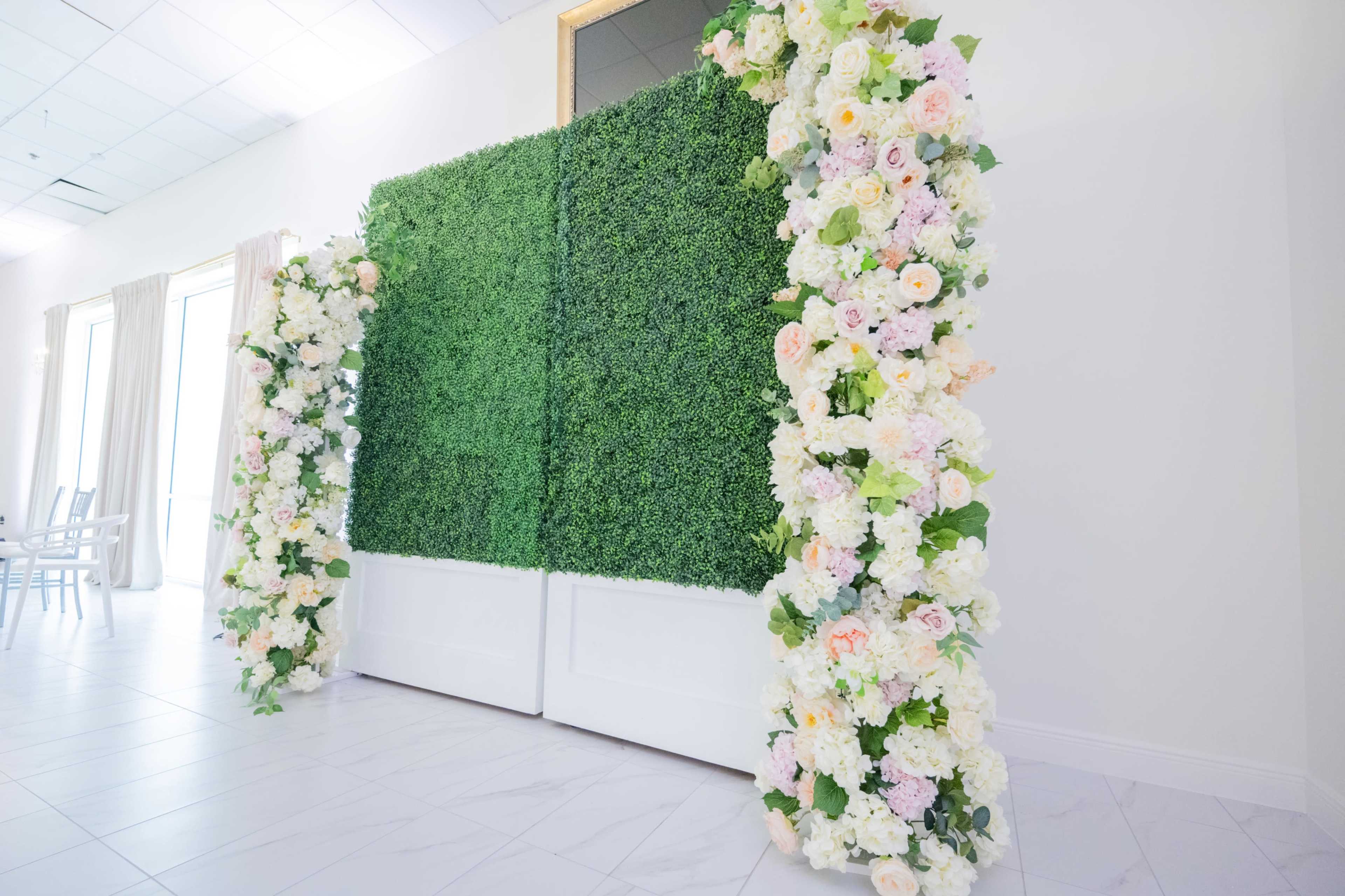 A greenery wall flanked by floral arrangements frames a pair of closed white doors in a bright interior space.