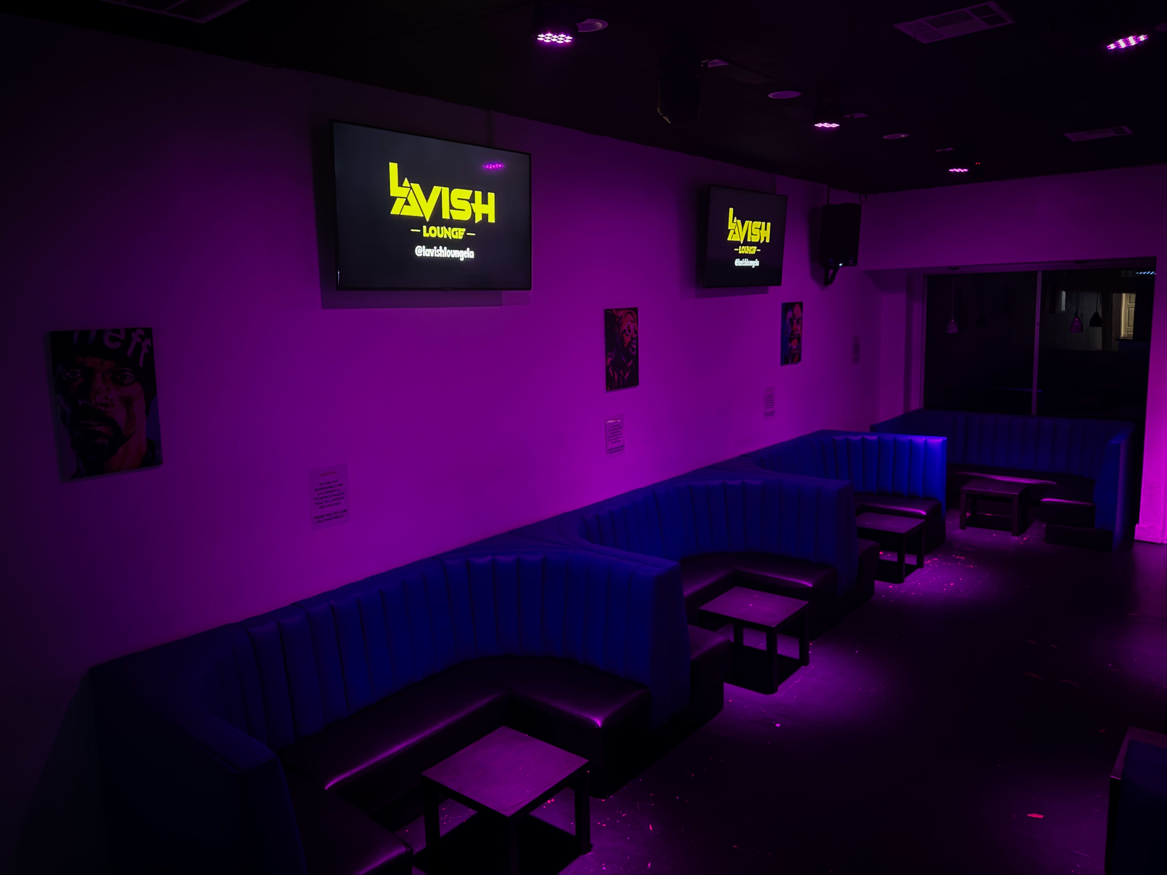 The image shows a dimly lit lounge area featuring purple lighting, blue seating, and two screens with the word "LAVISH" displayed.