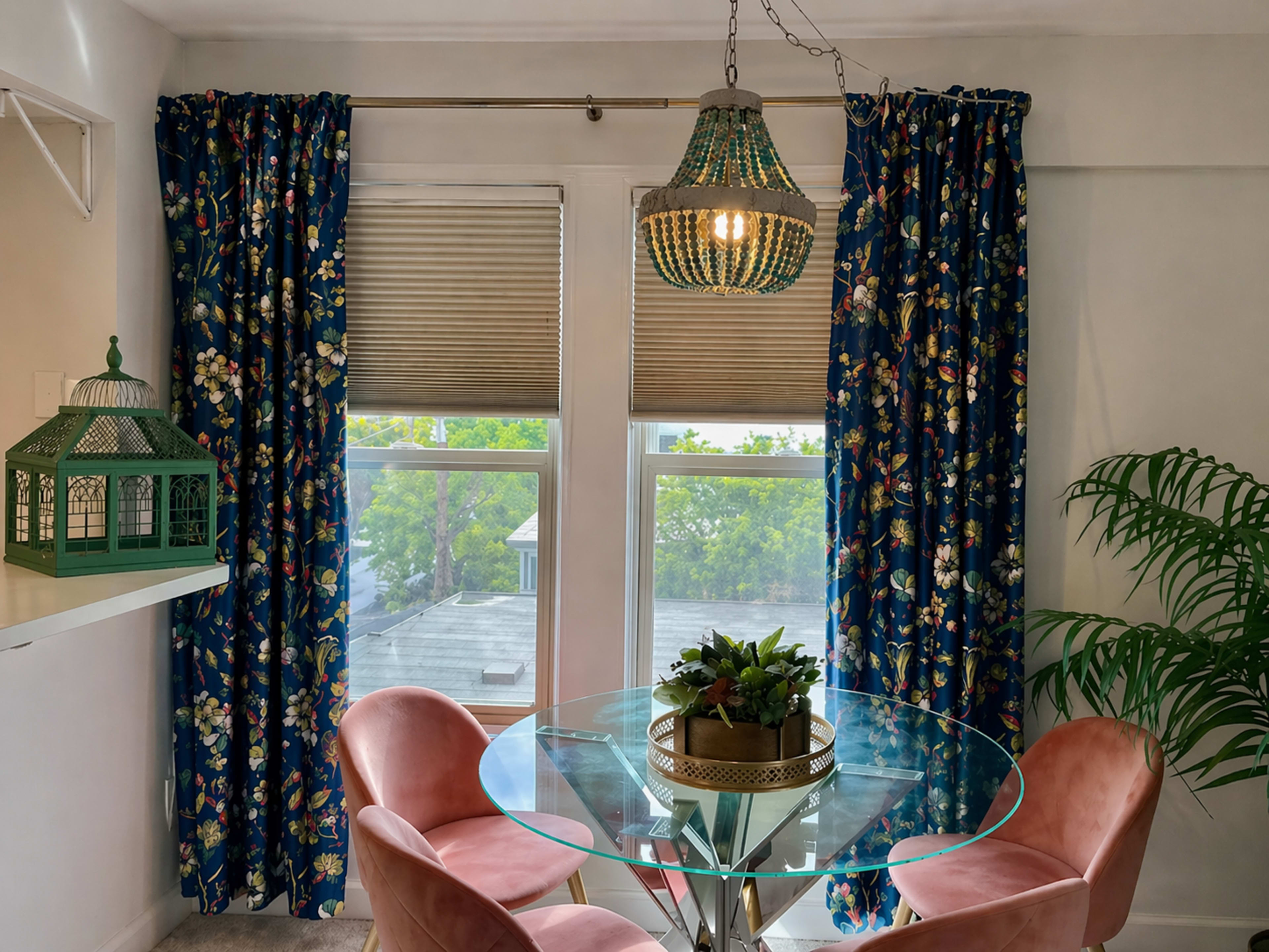 A round glass dining table with pink chairs is set near large windows dressed with floral curtains, while a green decorative lantern and a potted plant adorn the table.