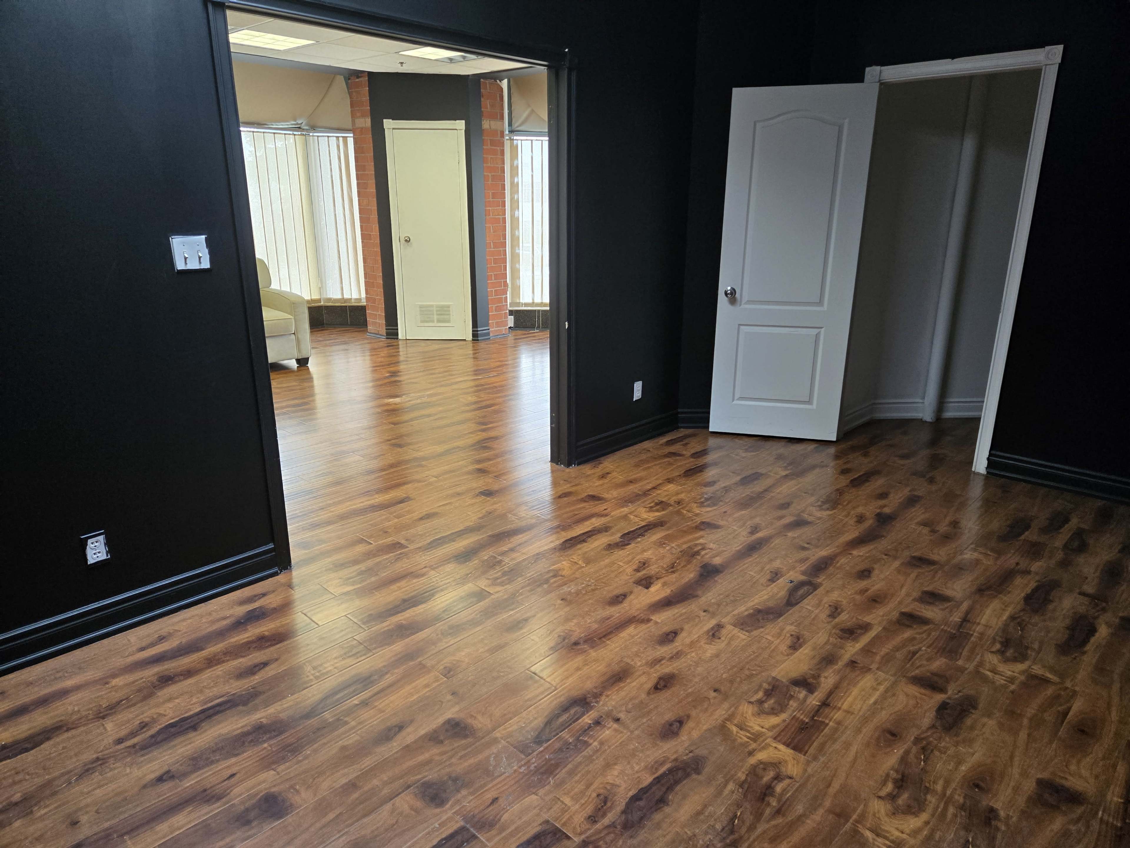 The image shows a partially furnished room with dark walls, wooden flooring, and an open doorway leading to another room with large windows.