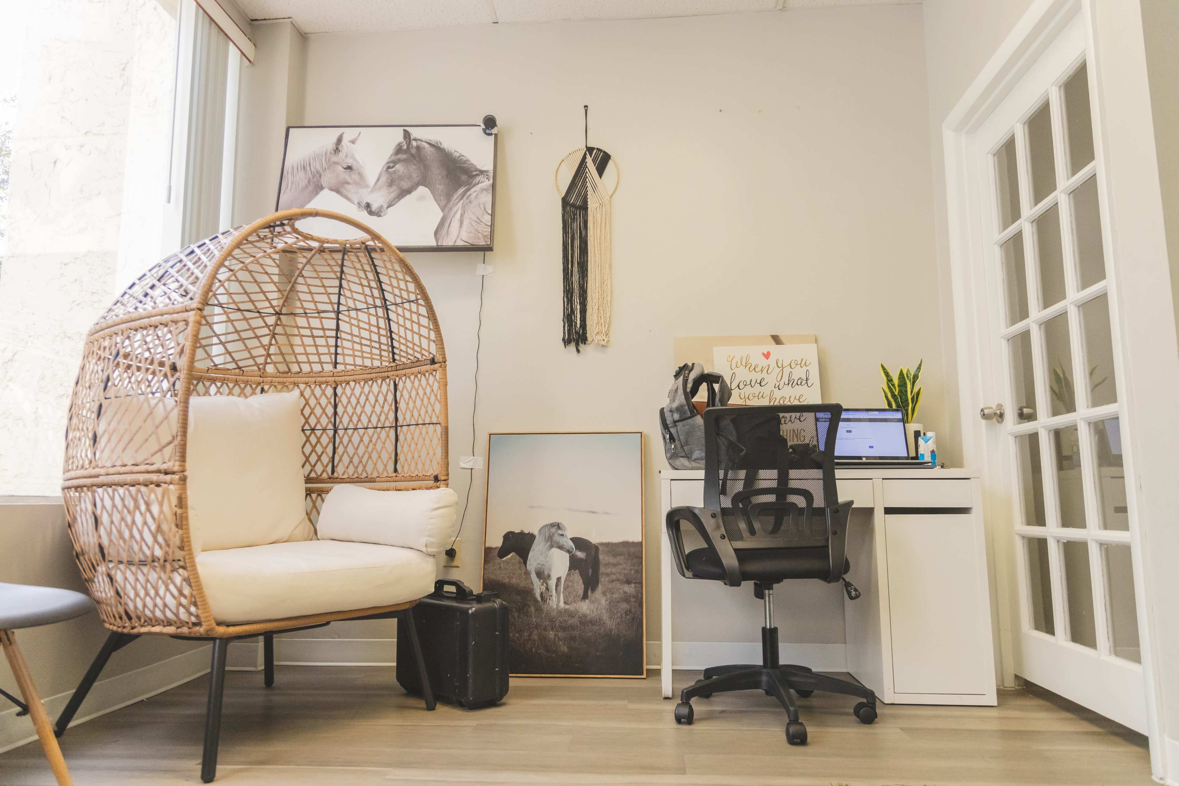 A cozy office space featuring a wicker chair, a small desk with a computer, and framed horse artwork on the walls.