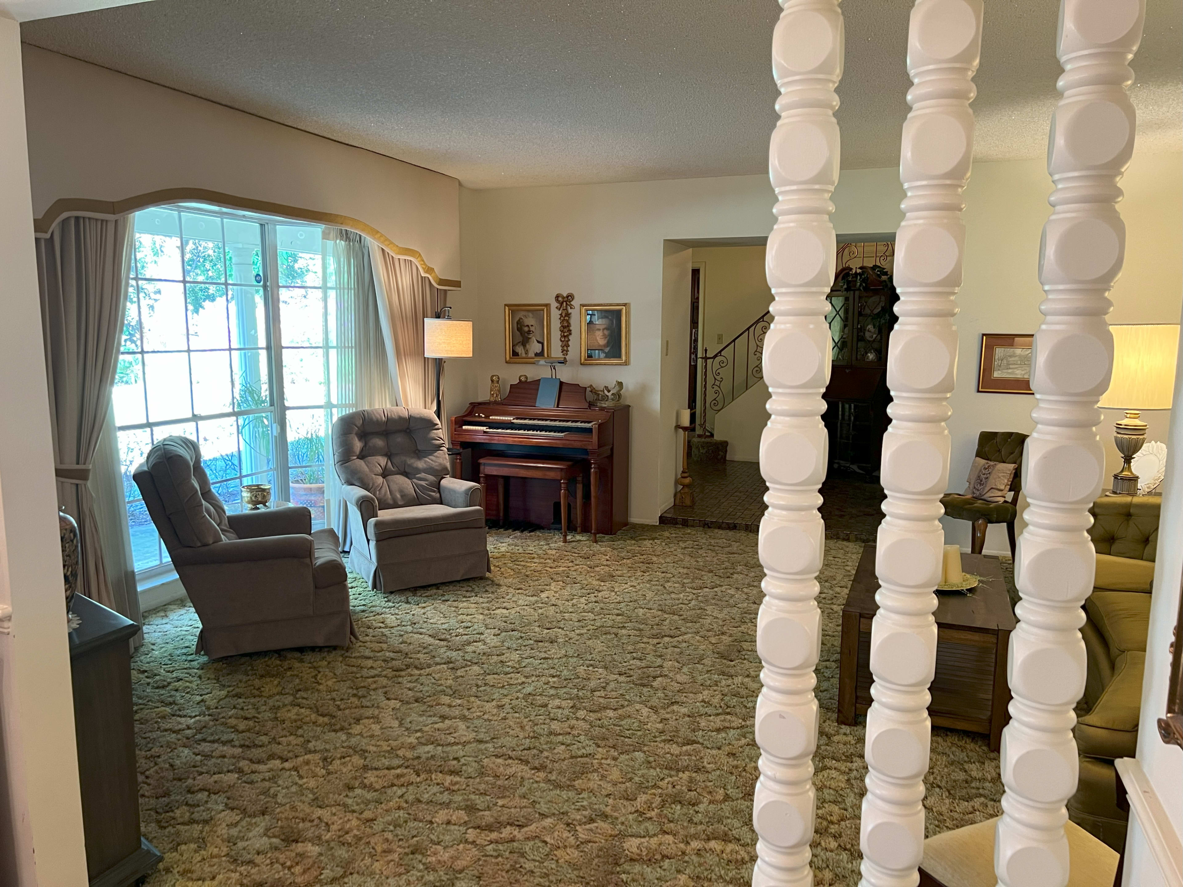 The image shows a spacious living room with two reclining chairs, a piano, and various decorative elements, all set on a carpeted floor.