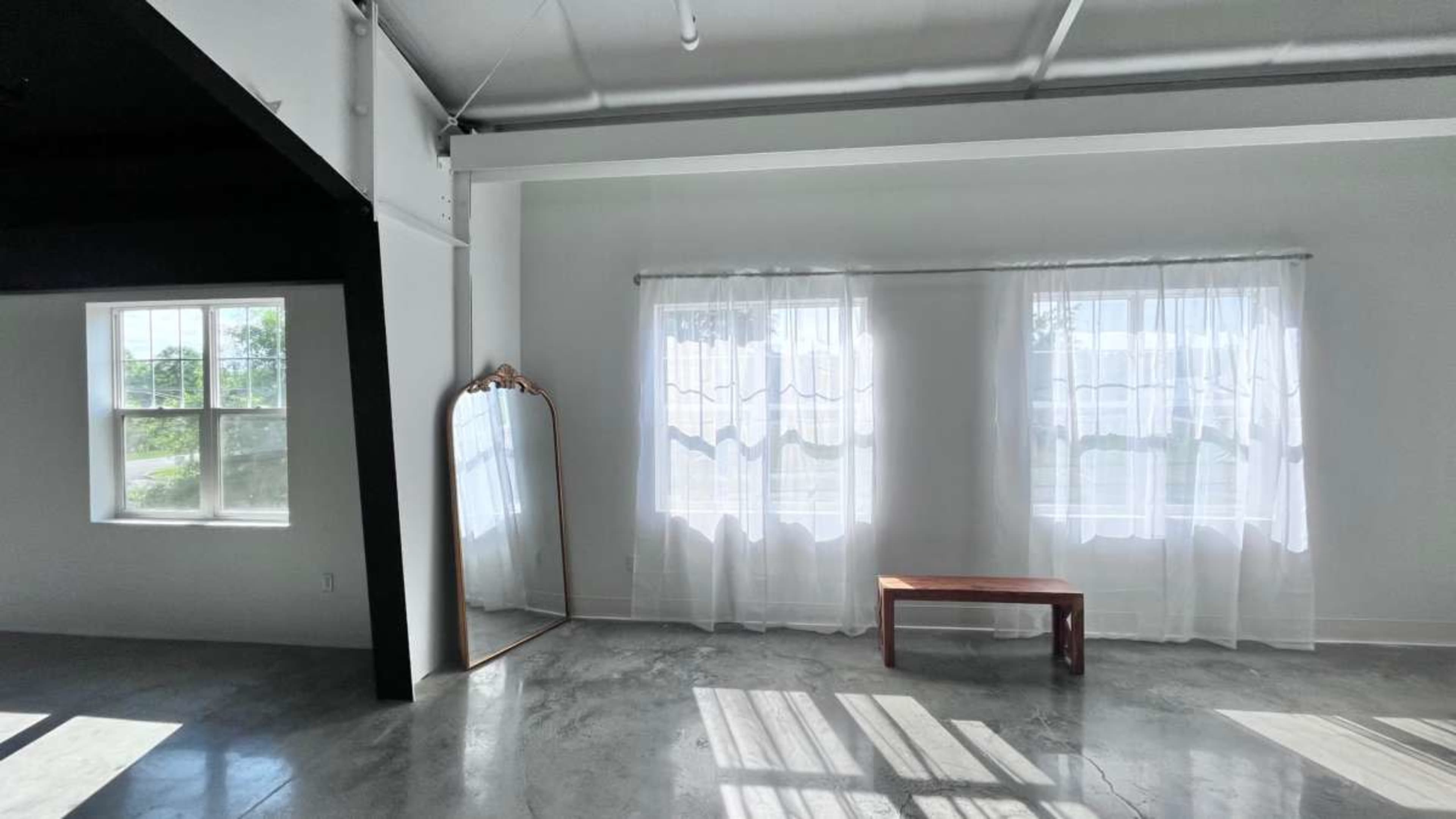 The image shows a bright, minimalistic interior with two large windows covered by sheer curtains and a wooden bench in front of a mirror against a white wall.