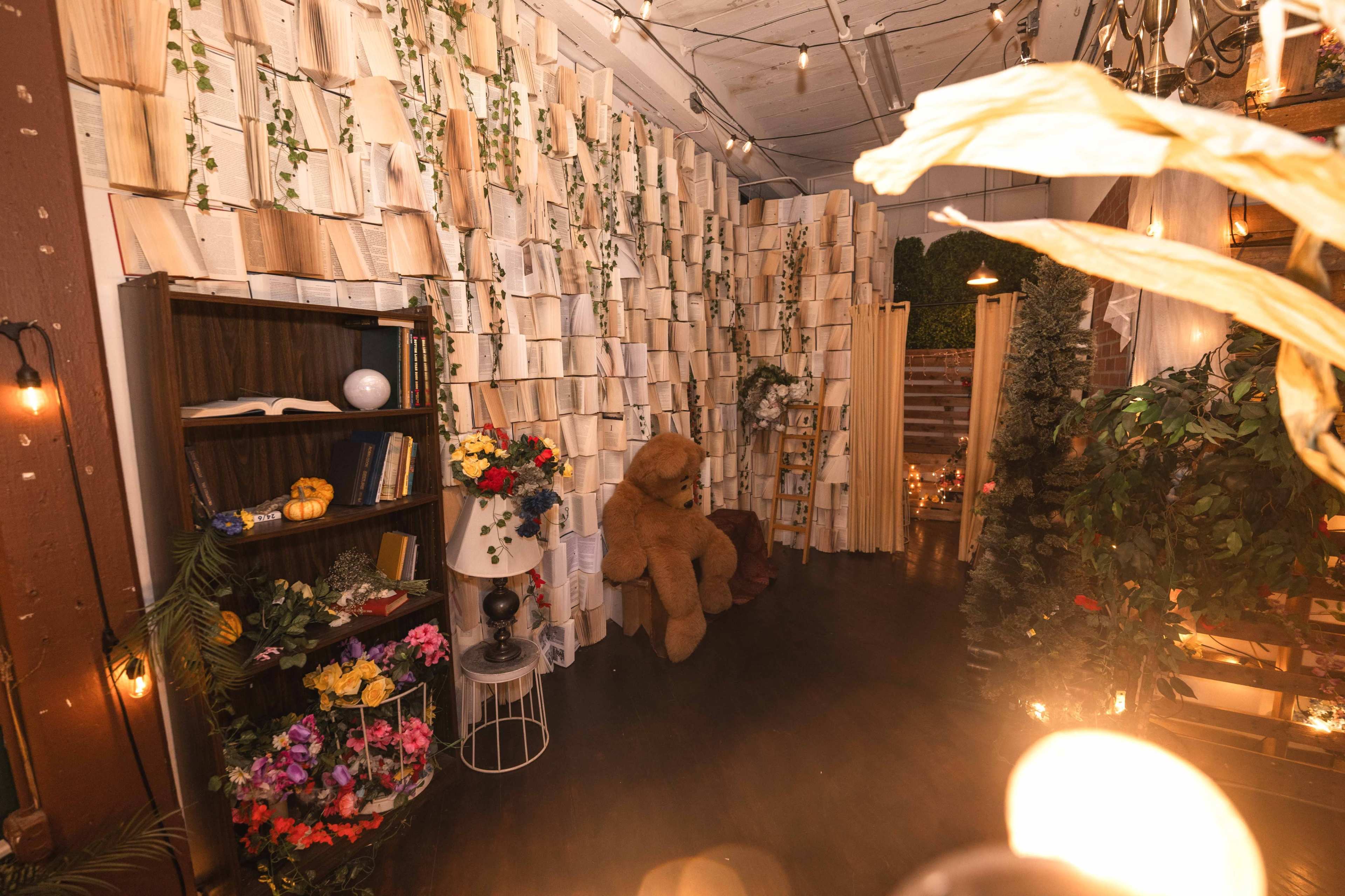 The image depicts a cozy indoor space featuring a wall of stacked books, a large teddy bear, a floral arrangement, and ambient lighting.
