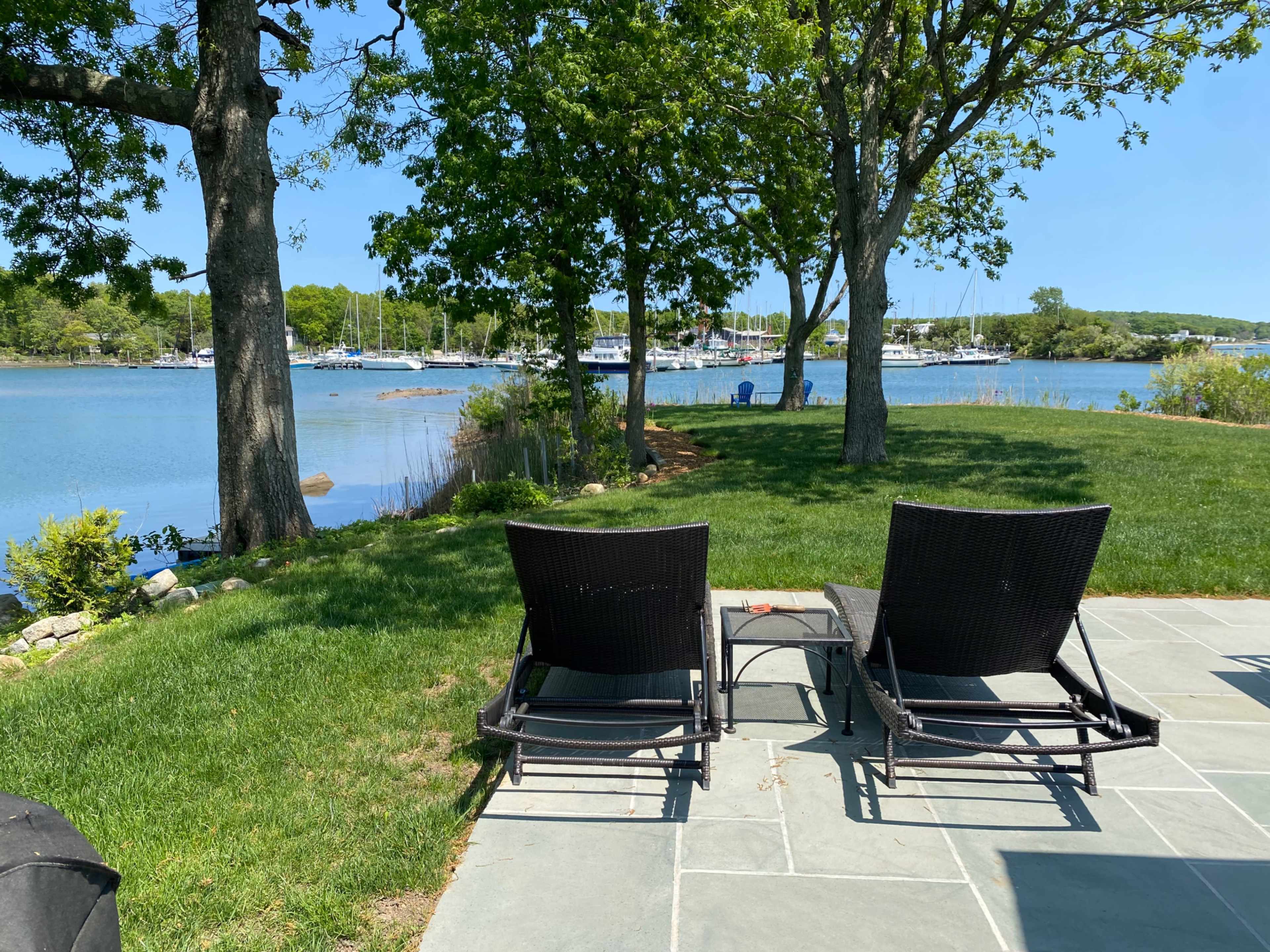 Two lawn chairs are positioned on a stone patio overlooking a calm waterway with boats in the distance, bordered by greenery and trees.