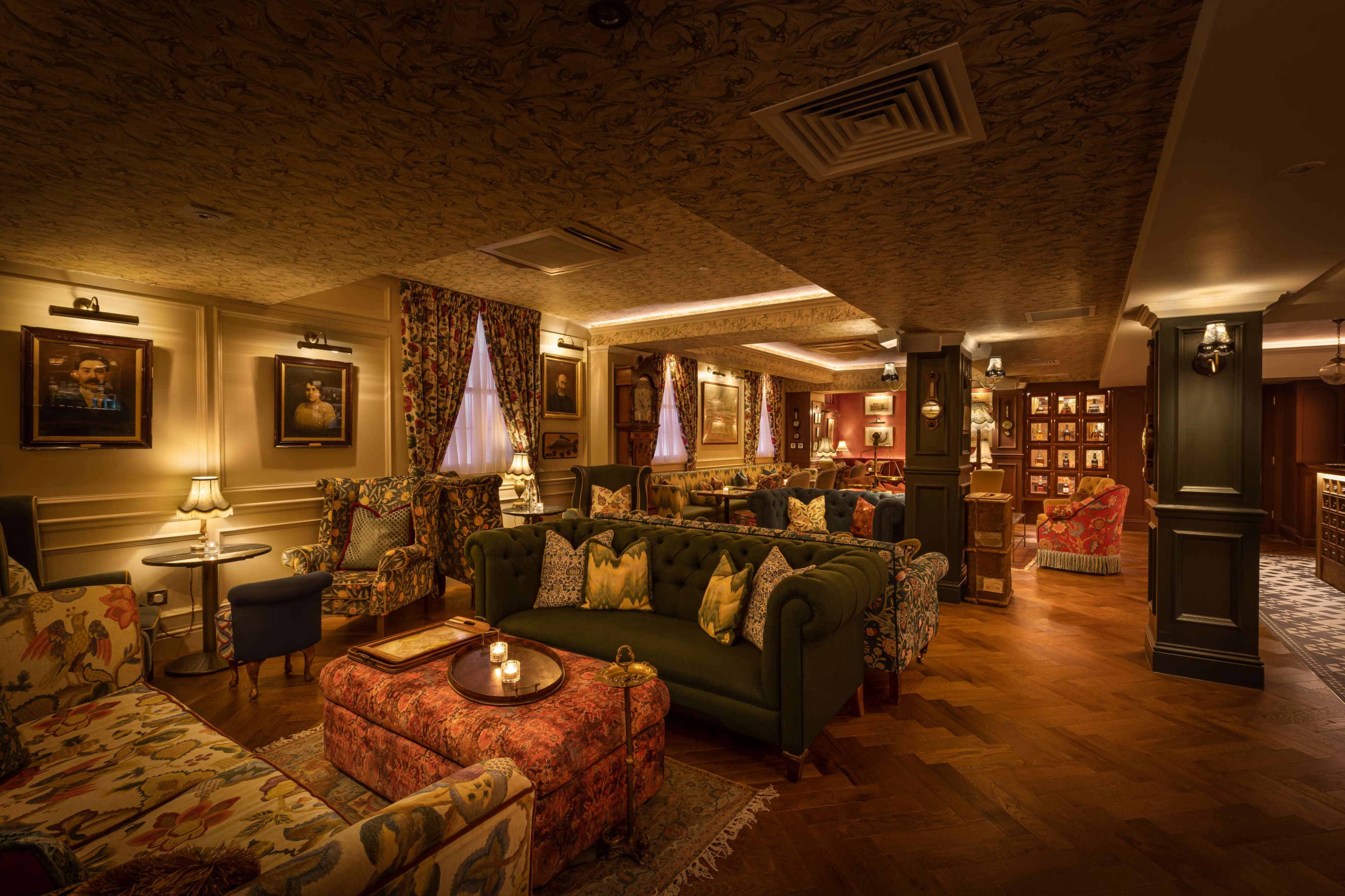 The interior of a cozy lounge features patterned sofas, floral curtains, and framed artwork on the walls, with warm lighting creating an inviting atmosphere.