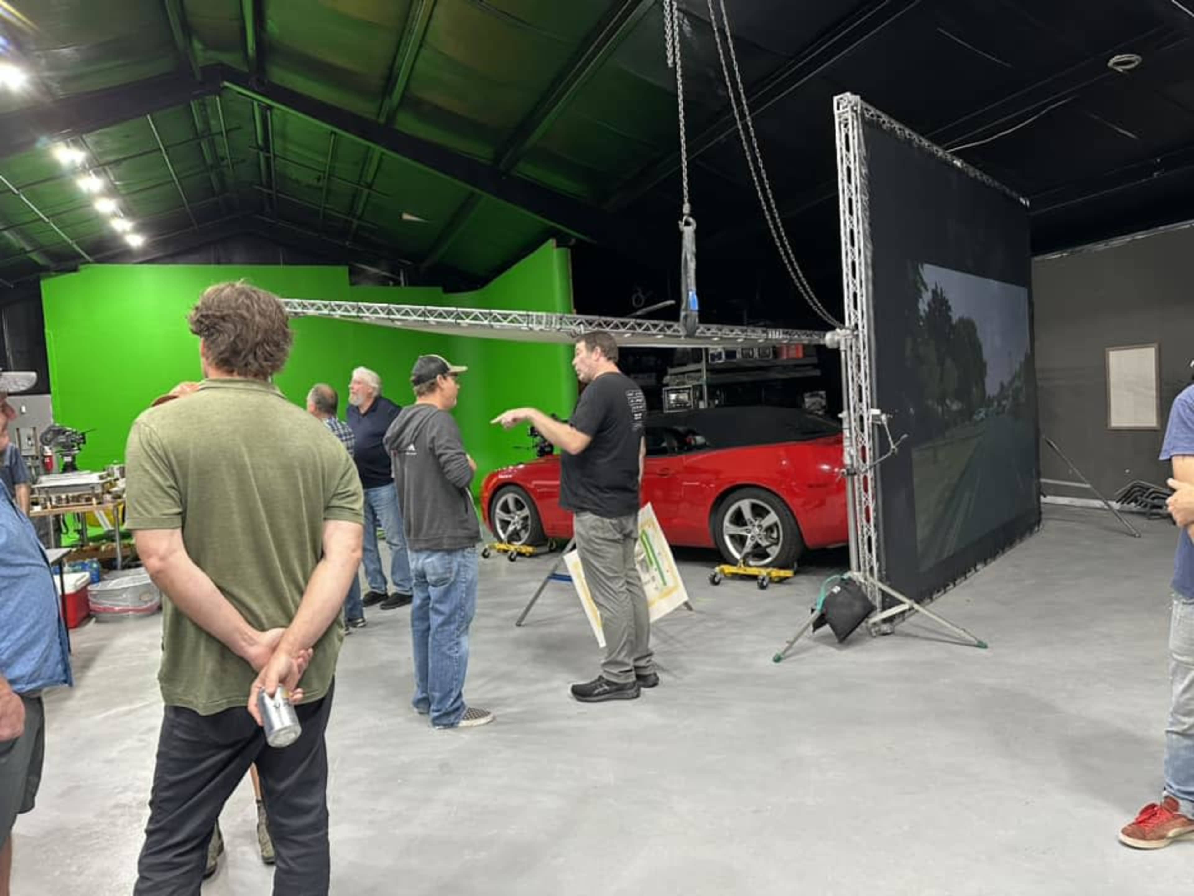 A group of people discusses in a studio with a red car and a large screen displaying a landscape, surrounded by green backdrop and studio equipment.