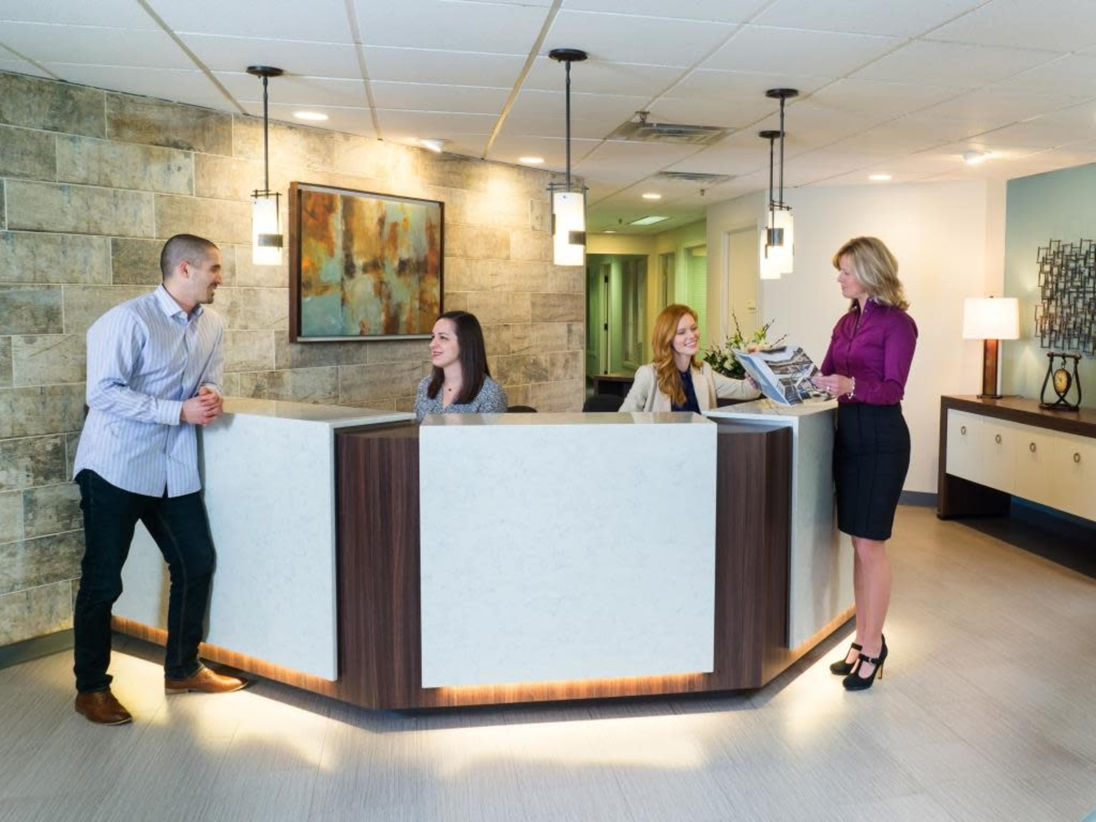 A modern reception area features a curved desk with three staff members interacting with a visitor, surrounded by neutral-toned walls and contemporary lighting.