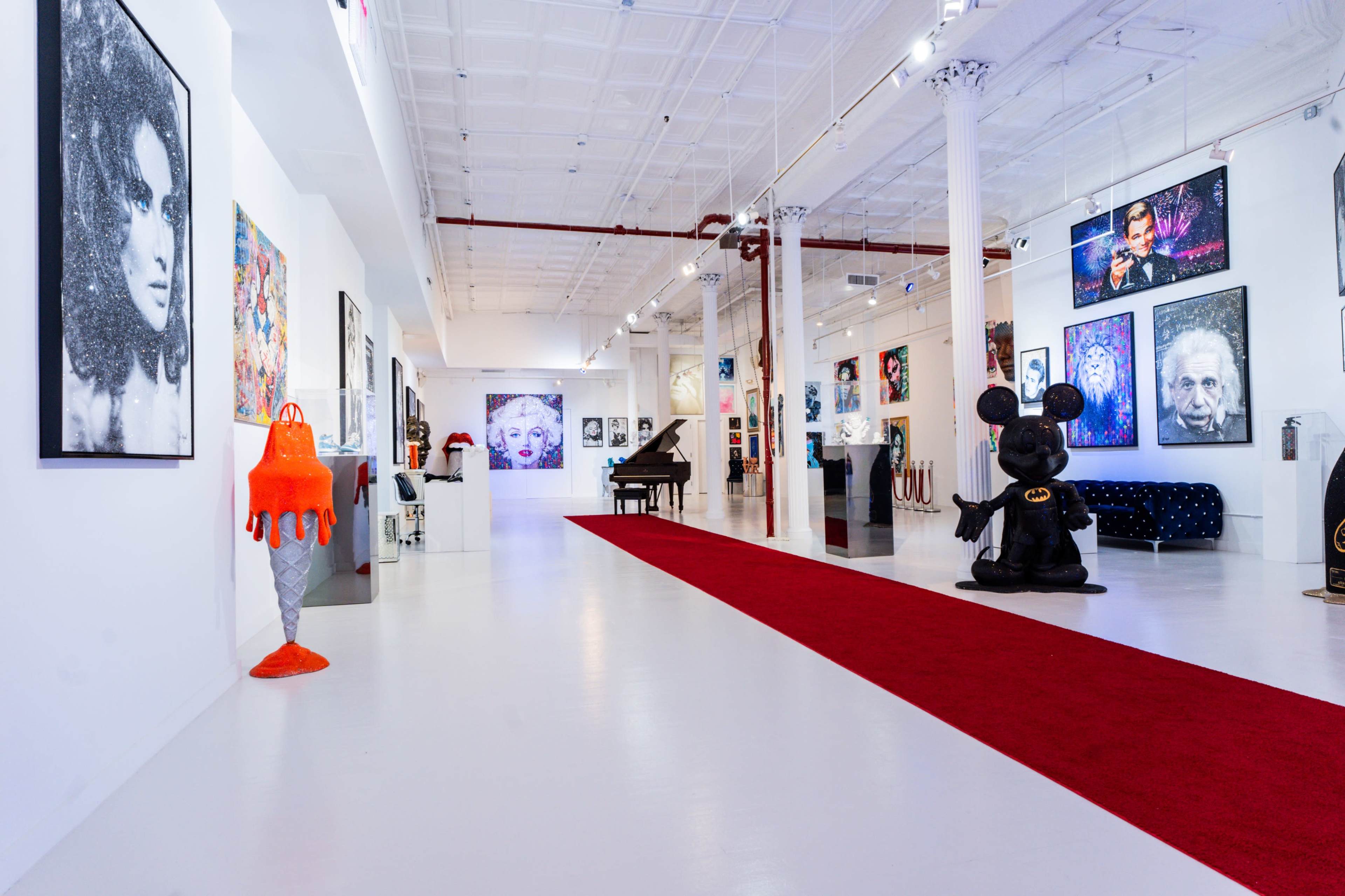 The image shows a spacious art gallery featuring various framed artworks on the white walls, a red carpet leading toward a grand piano, and a large black sculpture of a character in the foreground.