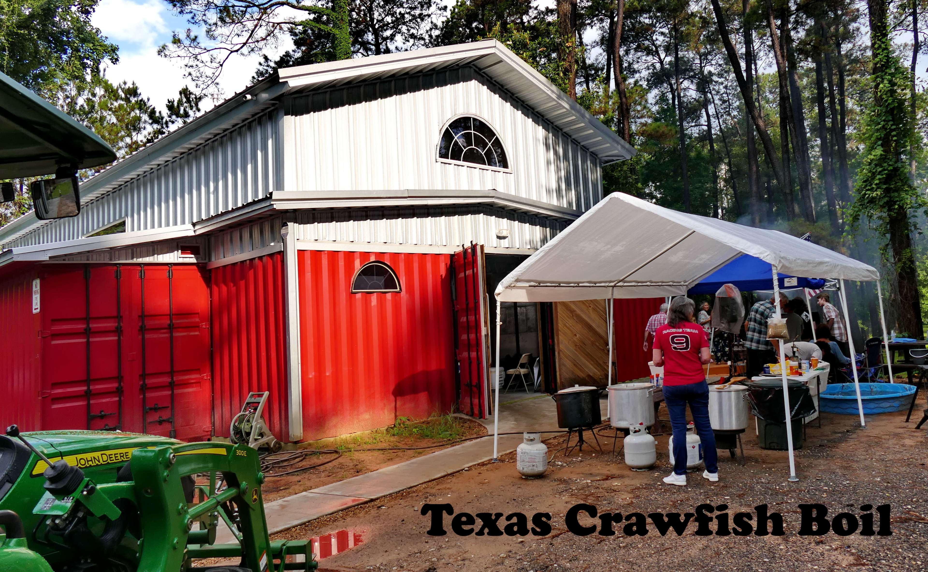 A person stands under a tent near a barn with red and silver exterior, preparing for a crawfish boil in a wooded area.