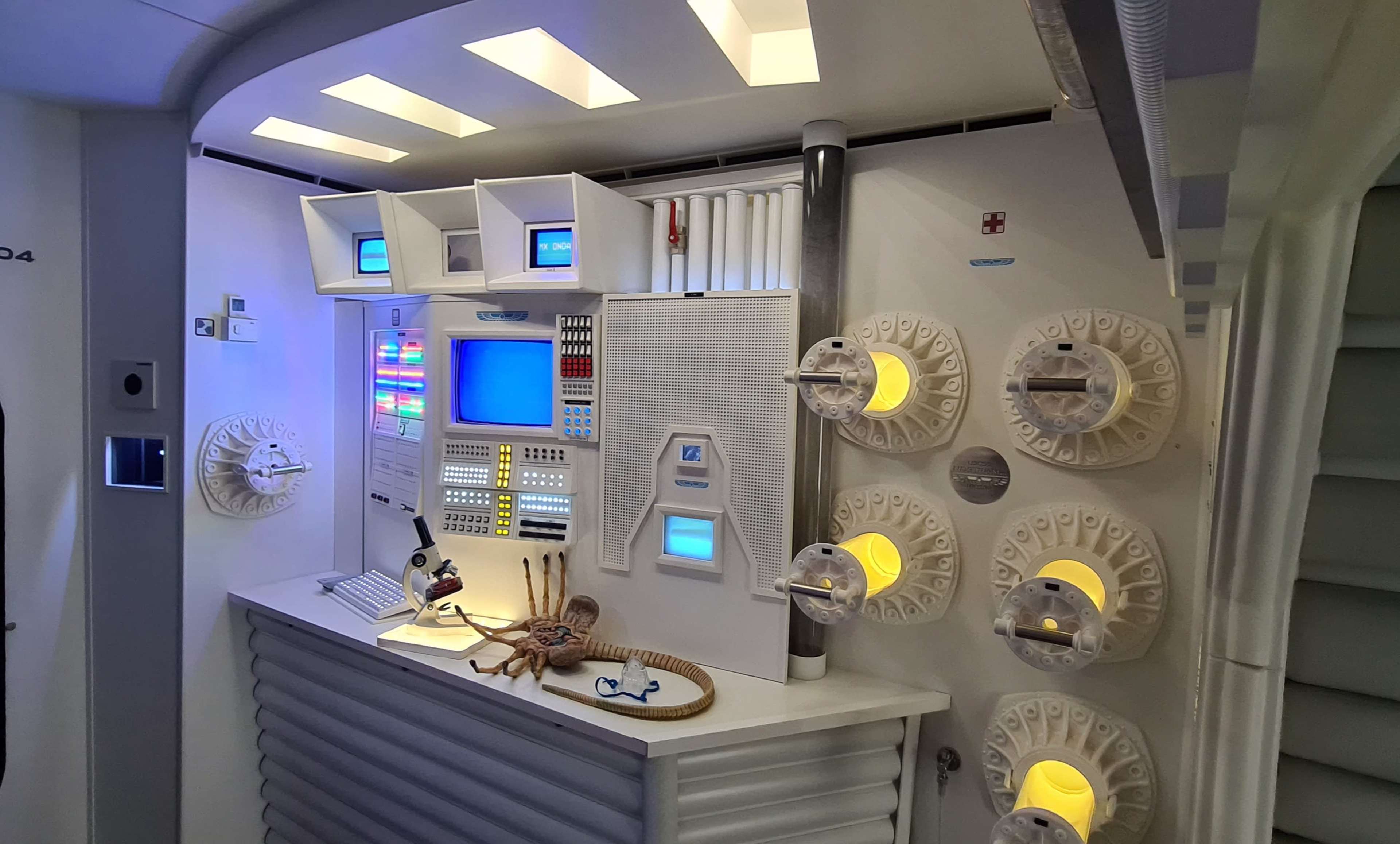 A futuristic control room with a white console featuring various screens, buttons, and circular panels along the walls.