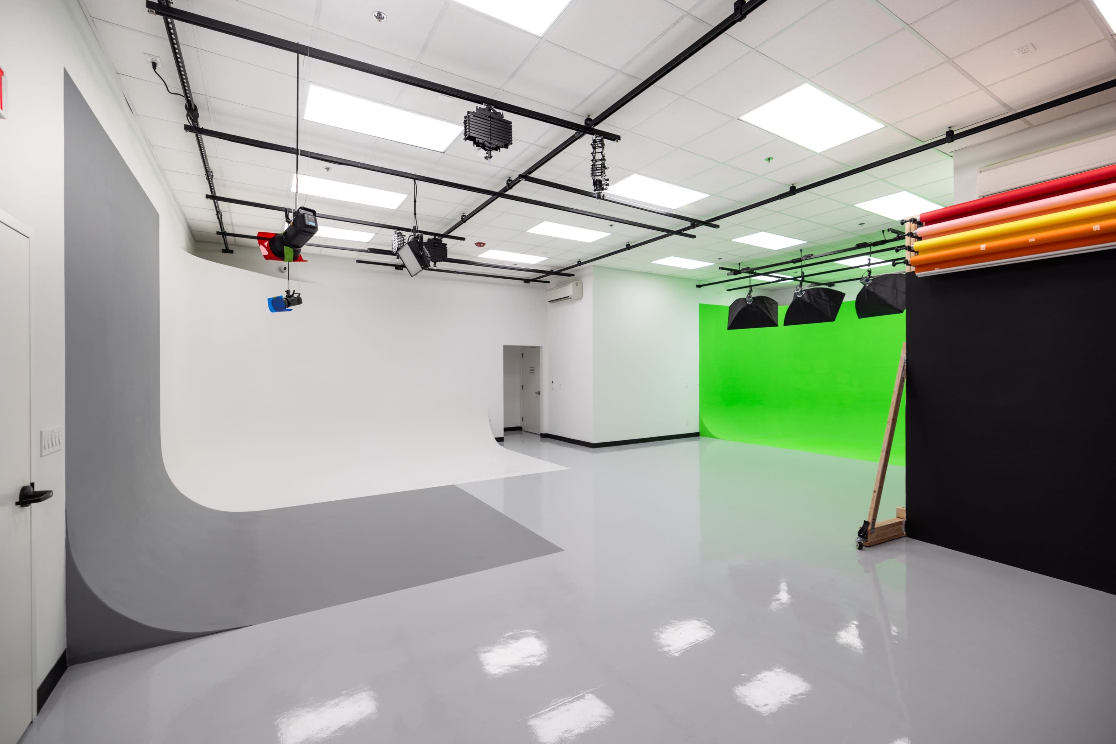 A spacious photo studio features a gray seamless backdrop on the left and a bright green screen on the right, with overhead lighting and a polished floor.