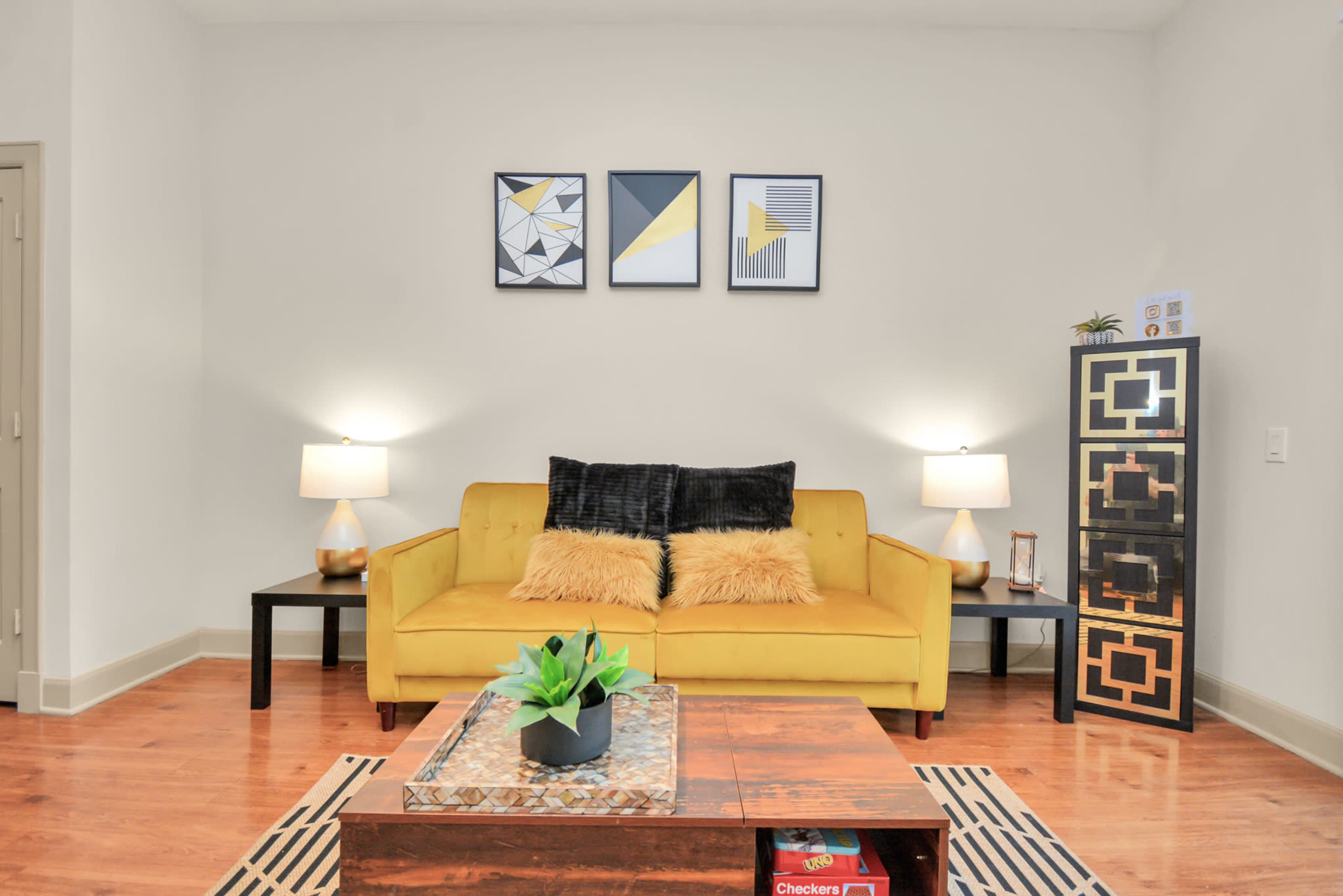 A yellow sofa with black and white cushions is positioned in a modern living room, accompanied by two lamps and a decorative side table.