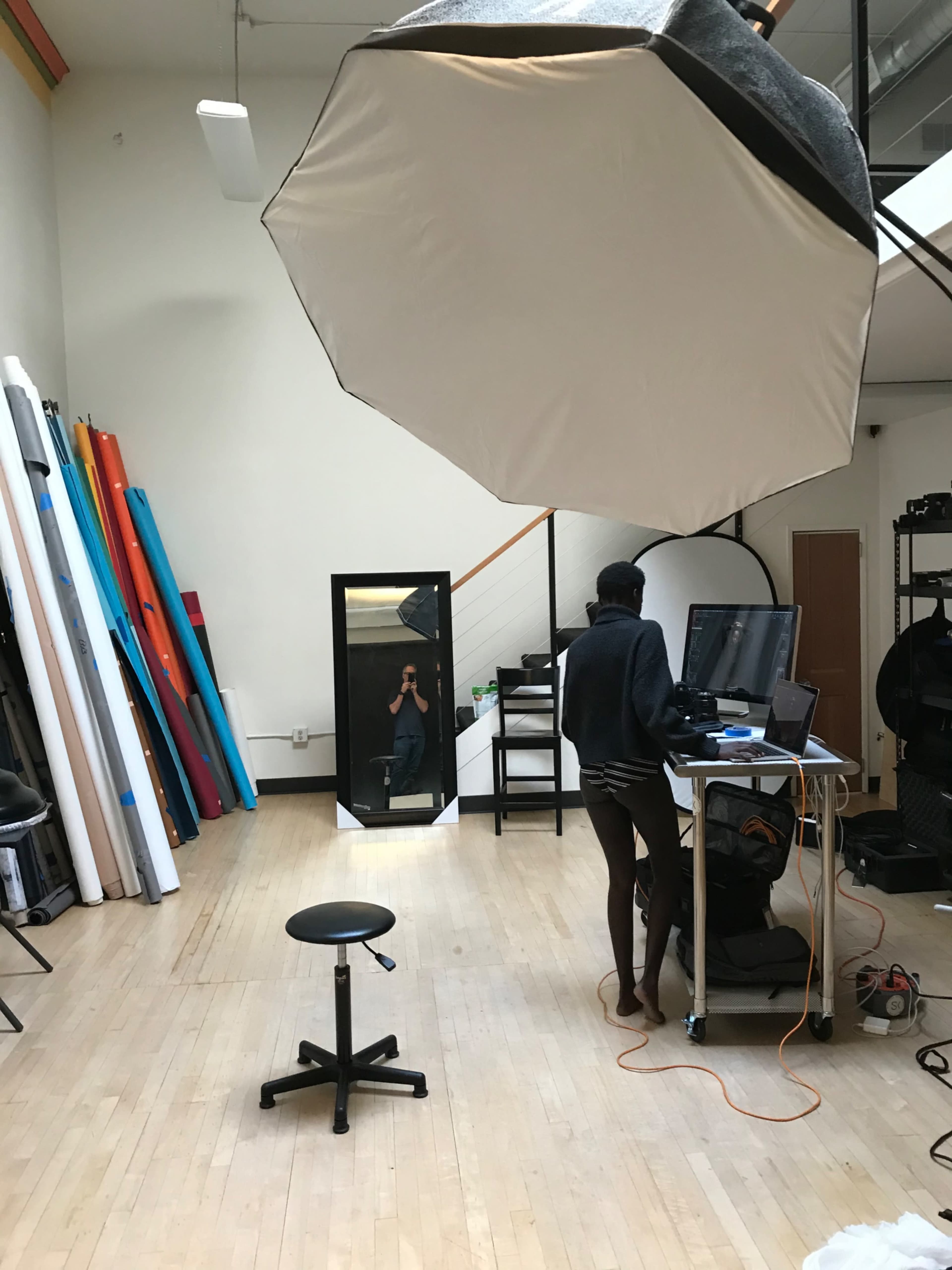 A photography studio with various colored backdrops, a large umbrella light, a mirror, and a person working at a computer.