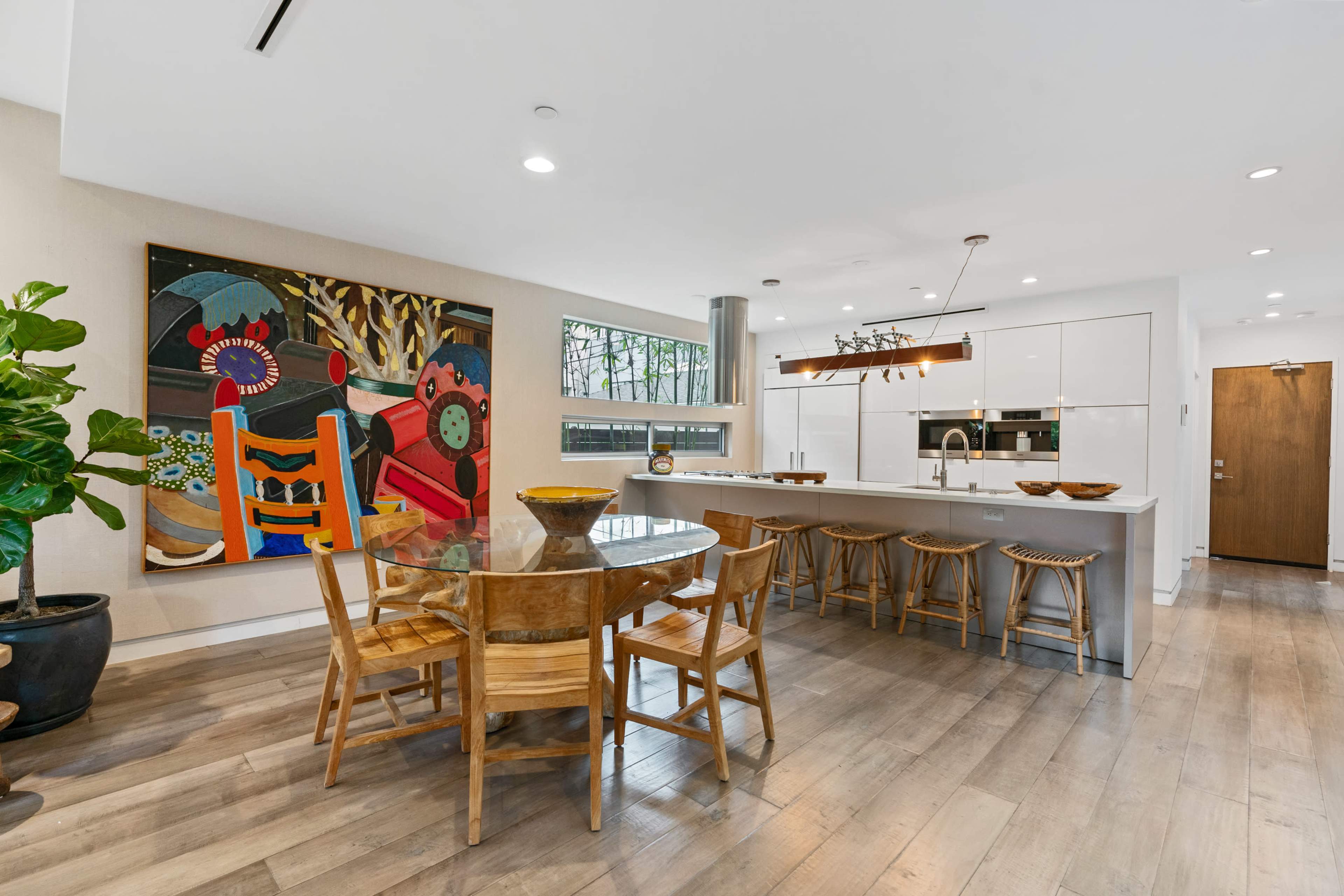 The image shows a modern kitchen and dining area featuring a round wooden table surrounded by chairs, a large colorful painting on the wall, and sleek cabinetry.