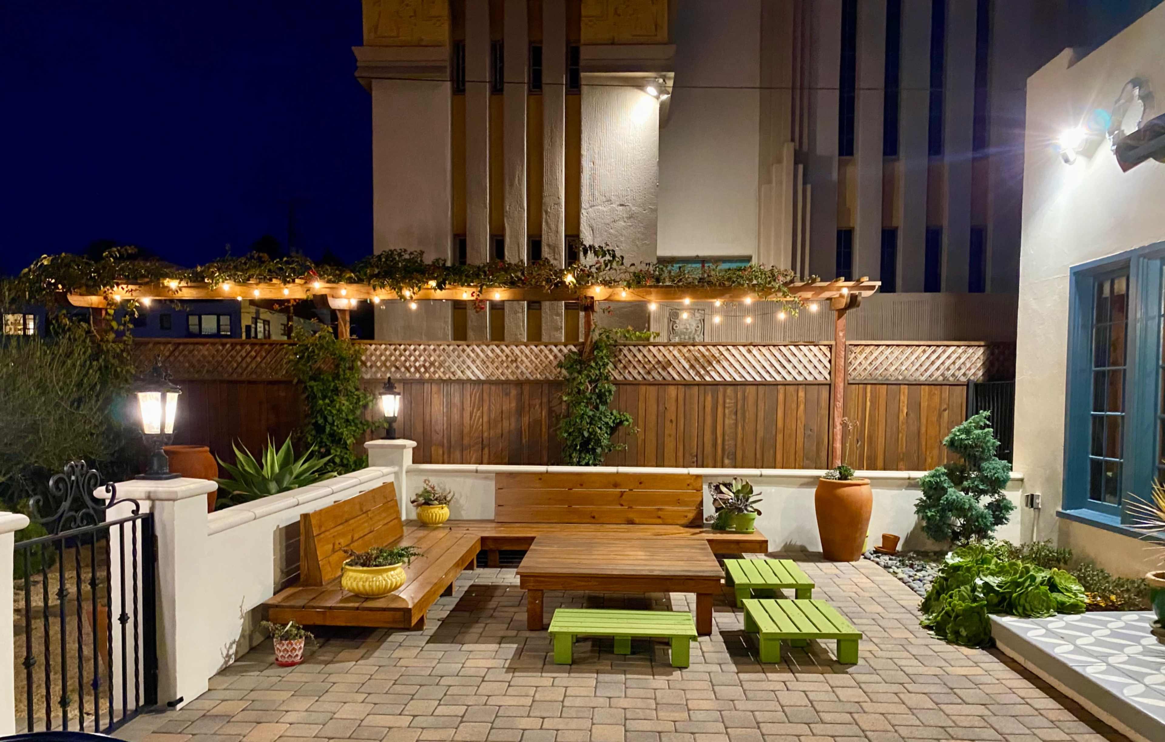 The image shows a well-lit outdoor patio area featuring wooden benches, green tables, and string lights overhead against a backdrop of a building with vertical stripes.