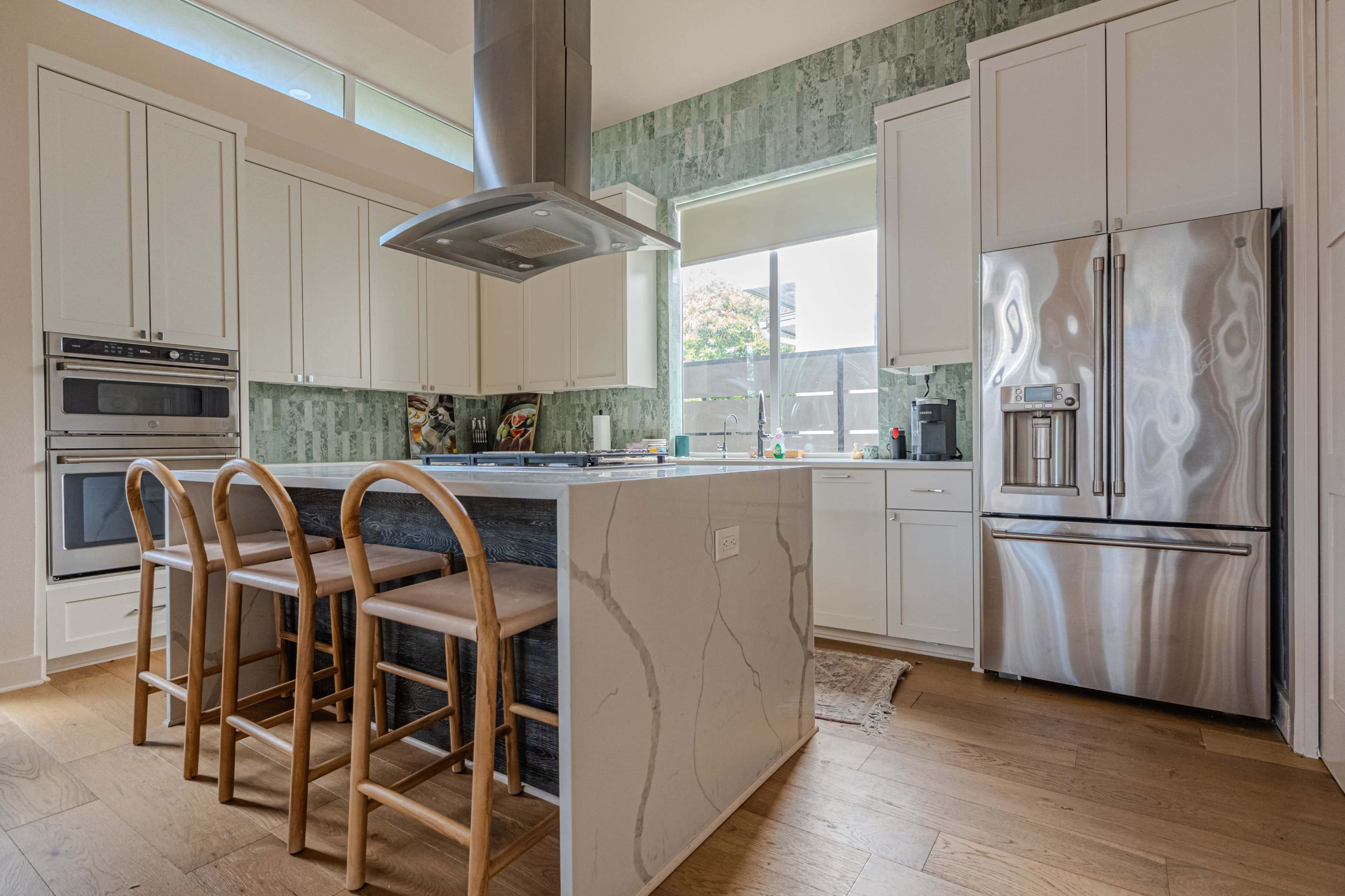 The image shows a modern kitchen with white cabinetry, stainless steel appliances, and a large island with wooden stools.