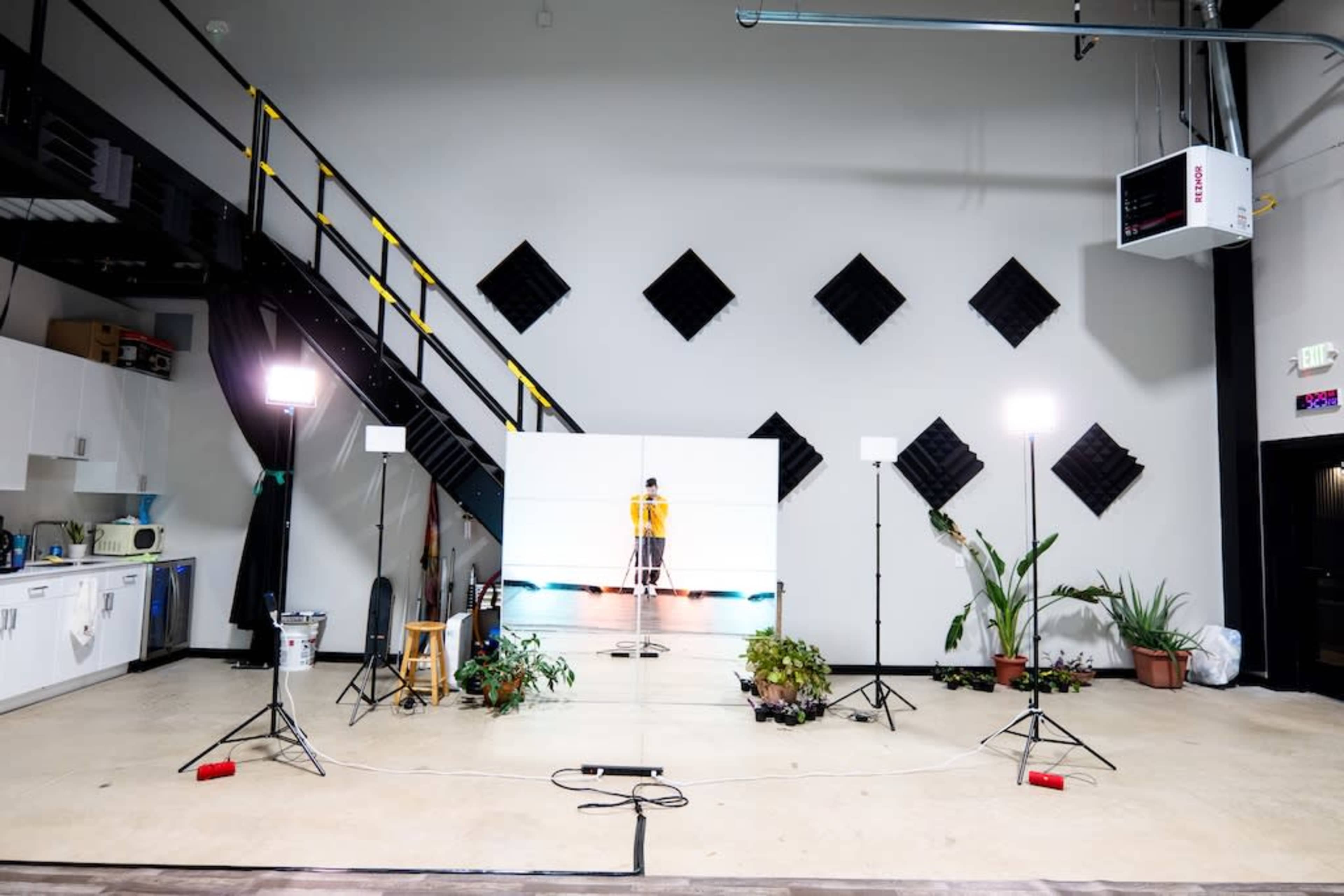 The image shows a studio setup with a central white backdrop, lighting equipment on either side, plants in pots, and a staircase in the background.