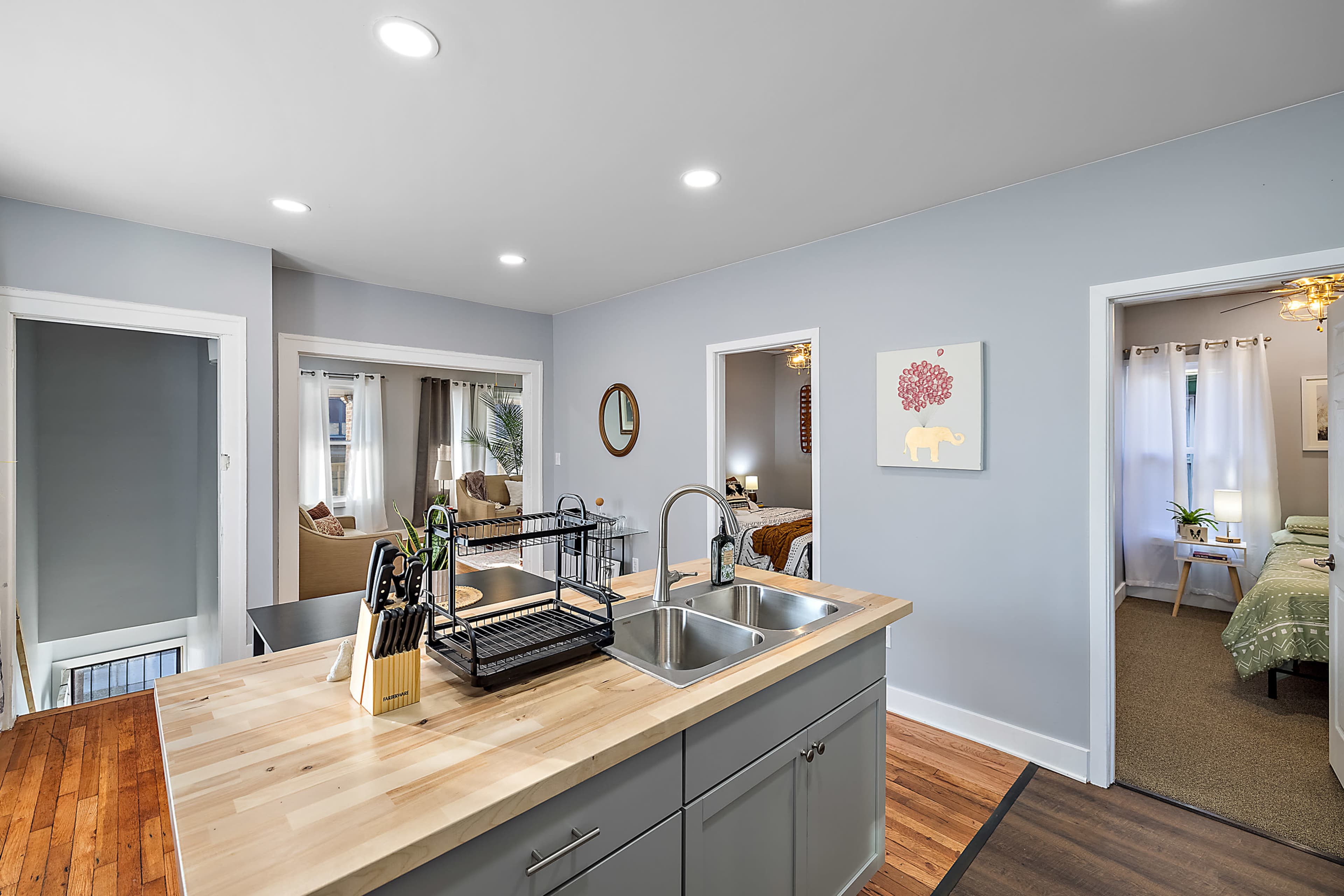 The image shows a modern kitchen area with a wooden island featuring a sink, leading to two doorways that open into a living room and a bedroom.