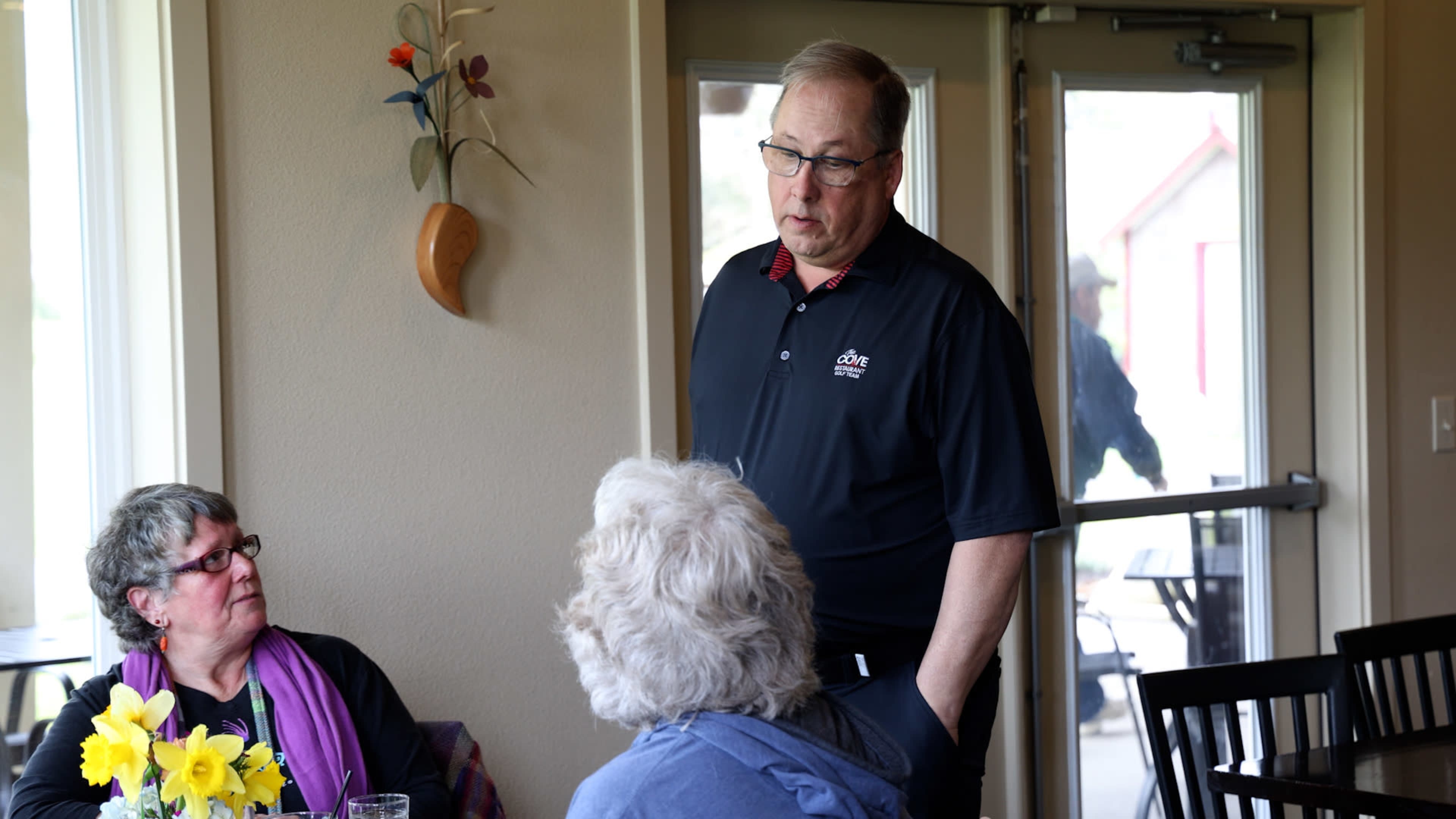 A man in a black polo shirt stands while speaking to two seated women in a dining area.