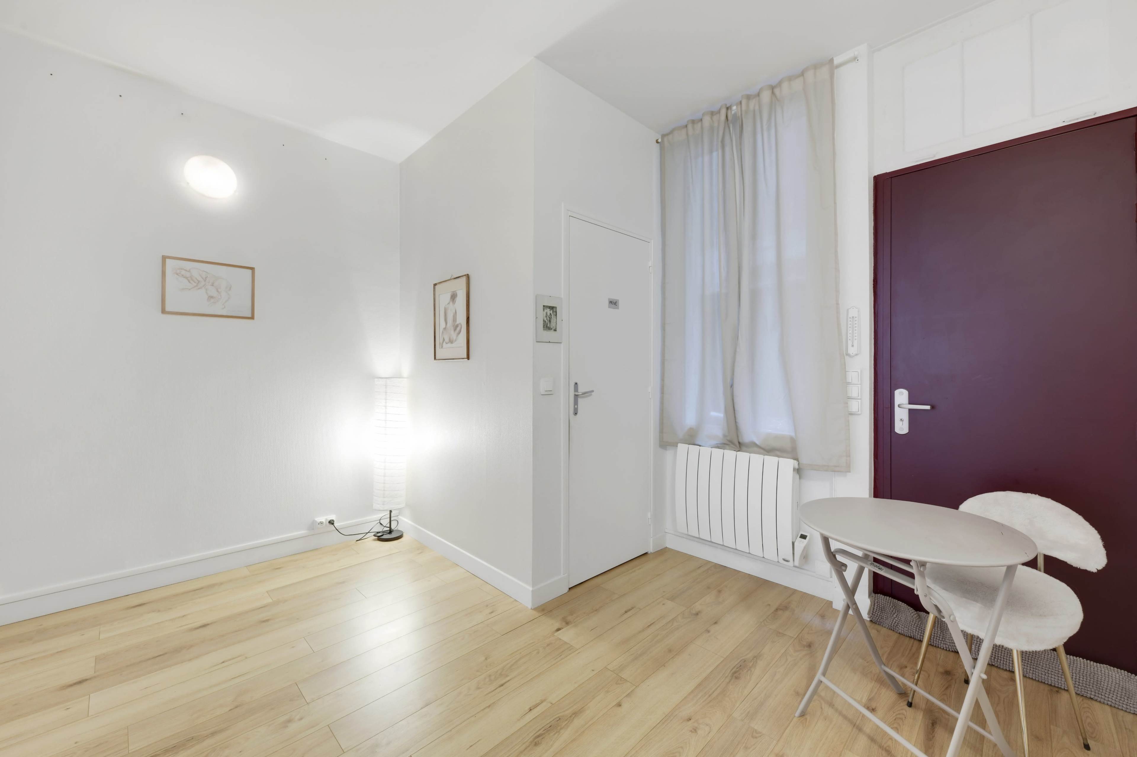 The image shows a small, minimalist room featuring a table and two chairs, a standing lamp, a door with a maroon panel, and a window draped with a sheer curtain.