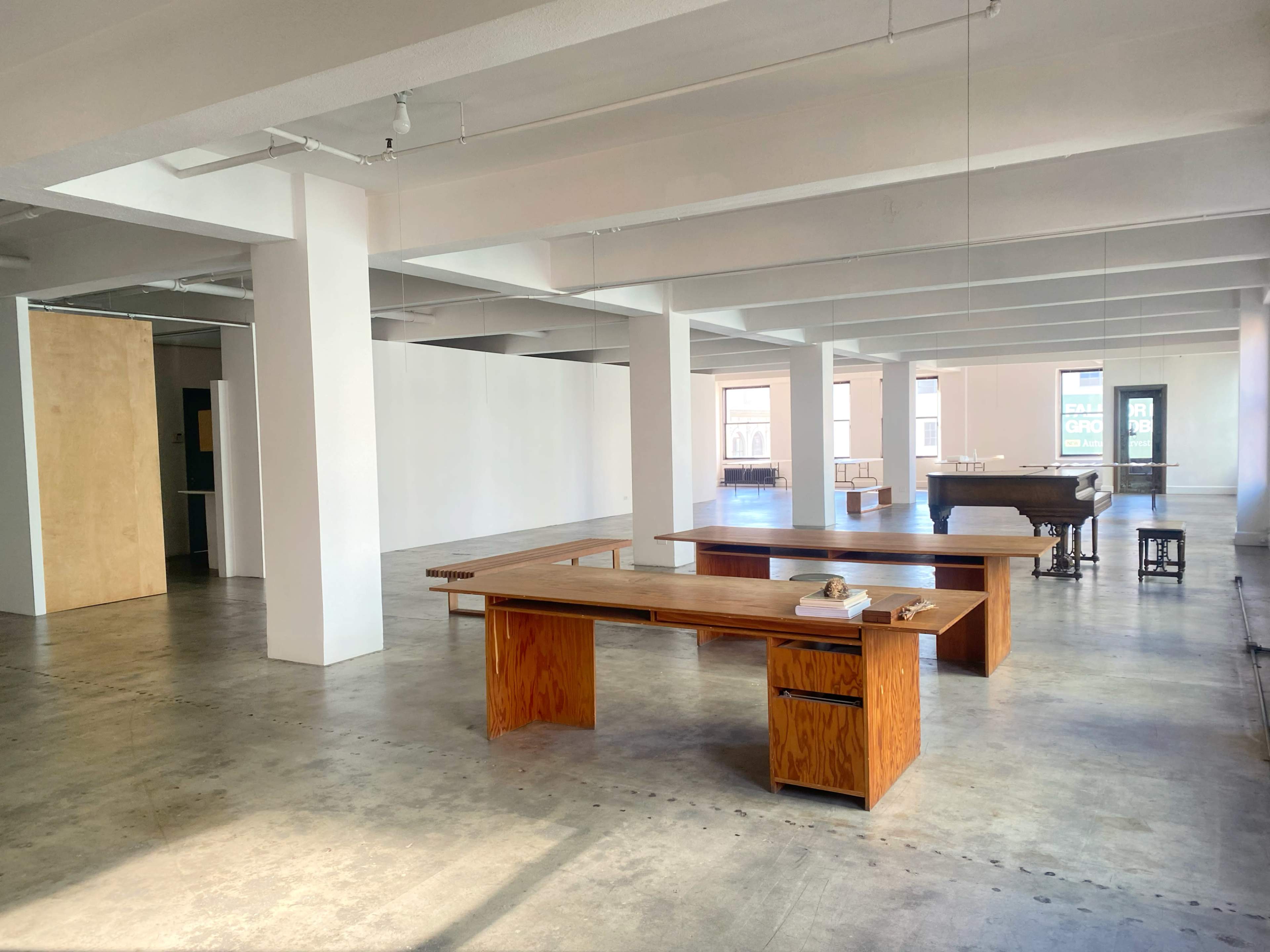 The image shows a spacious interior of a modern open area with wooden furniture, including tables, and a piano, surrounded by plain white walls and polished concrete floors.