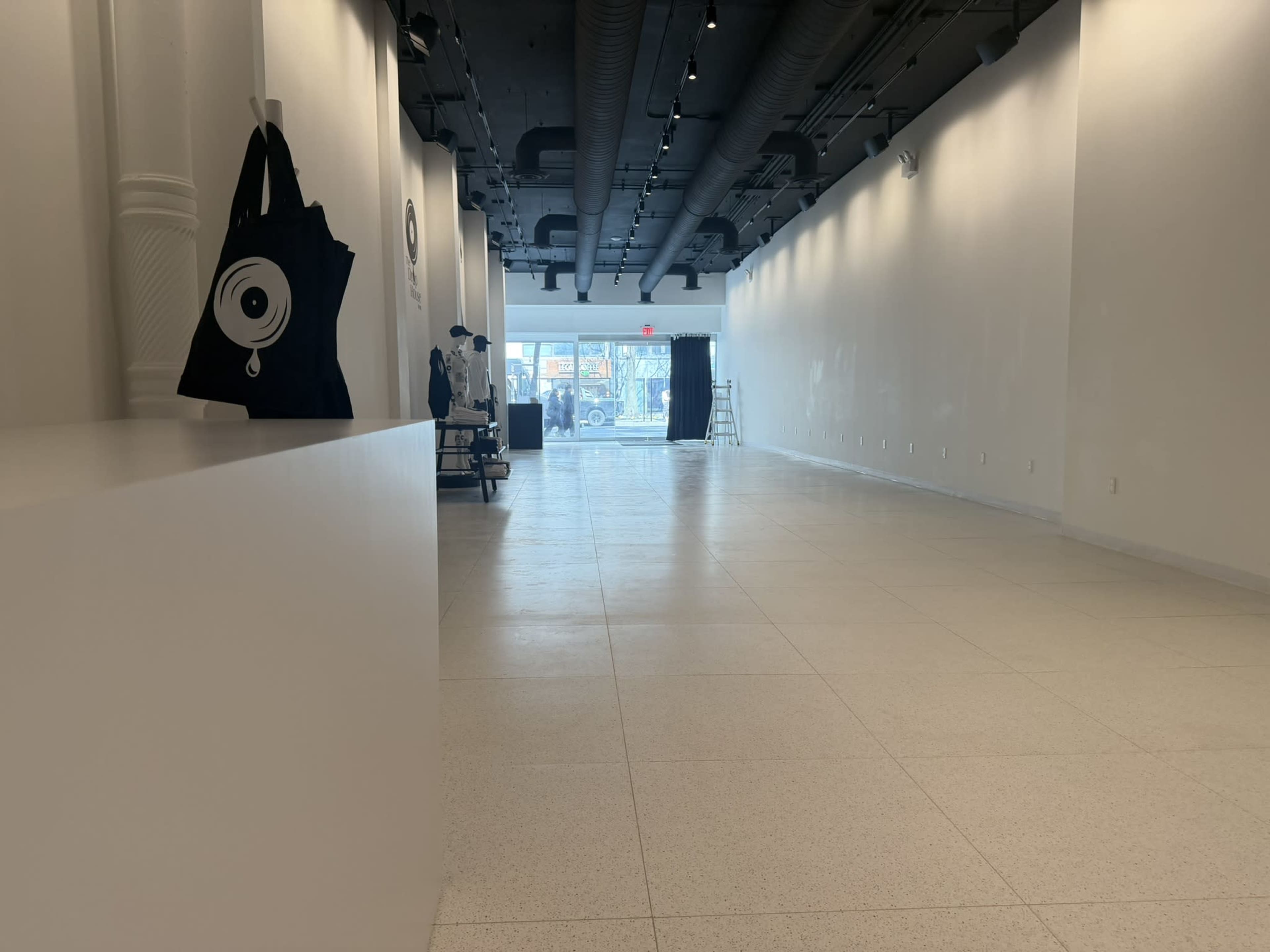 The image shows a spacious, minimally decorated retail space with white walls and a lightly textured floor, featuring a black tote bag on a white ledge and a view of the street through a large front window.