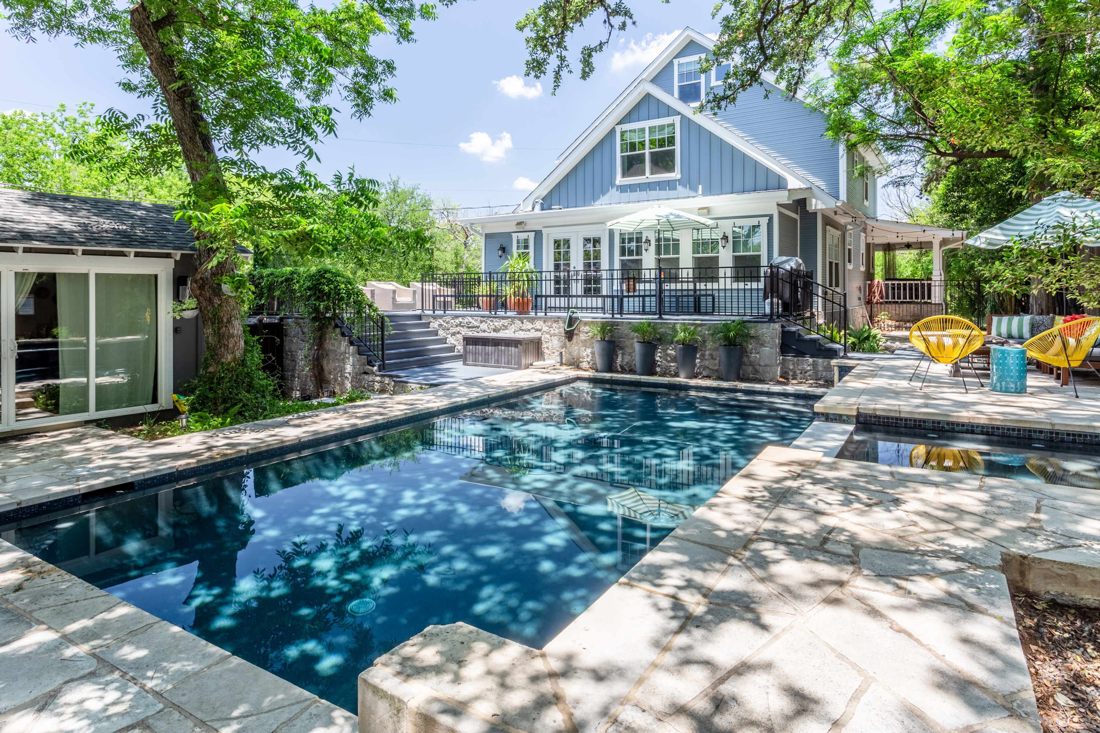 The image shows a rectangular swimming pool surrounded by stone decking and a landscaped yard with trees and outdoor furniture, adjacent to a large blue house.