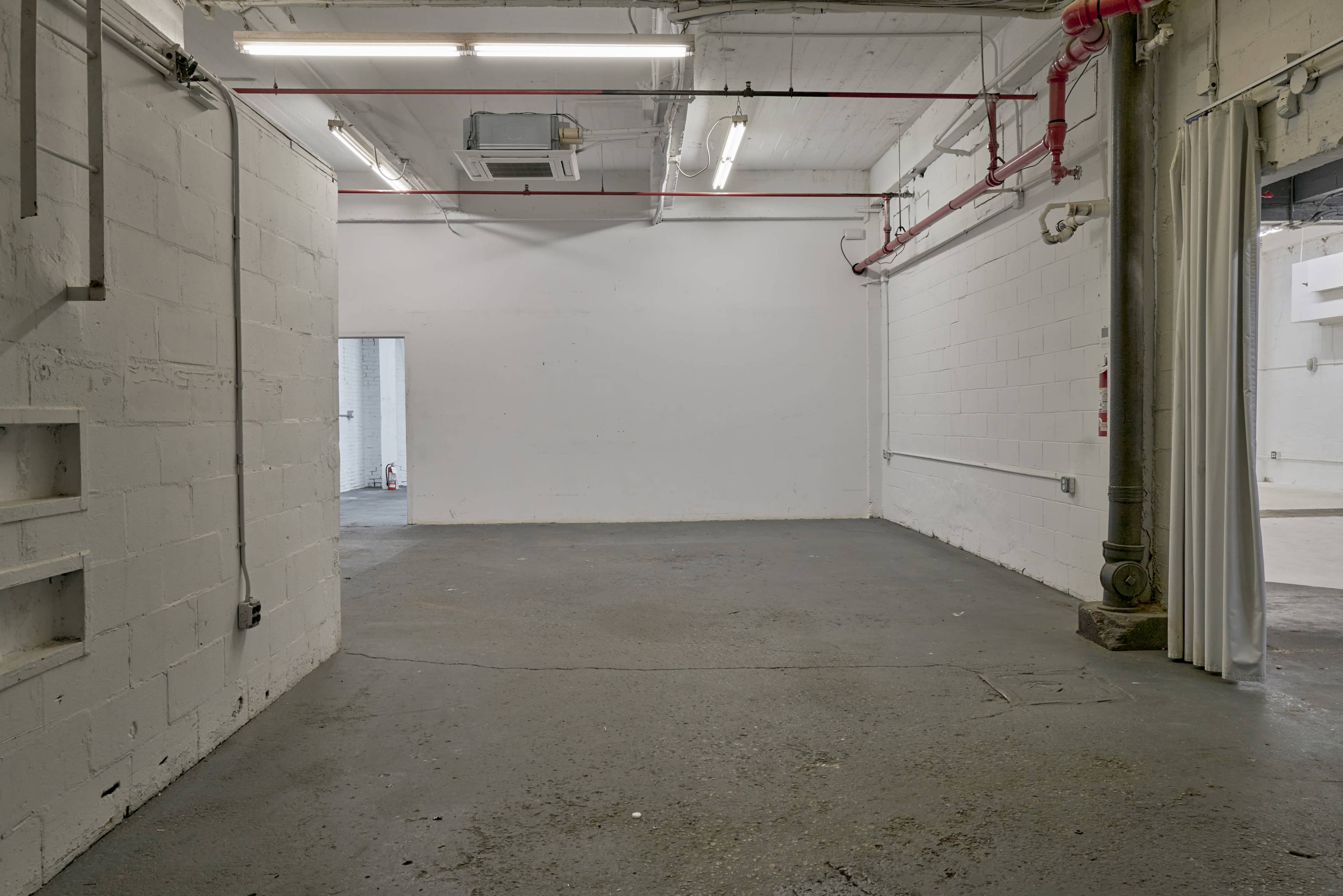 The image shows an empty industrial space with white walls, concrete flooring, and exposed pipes along the ceiling.