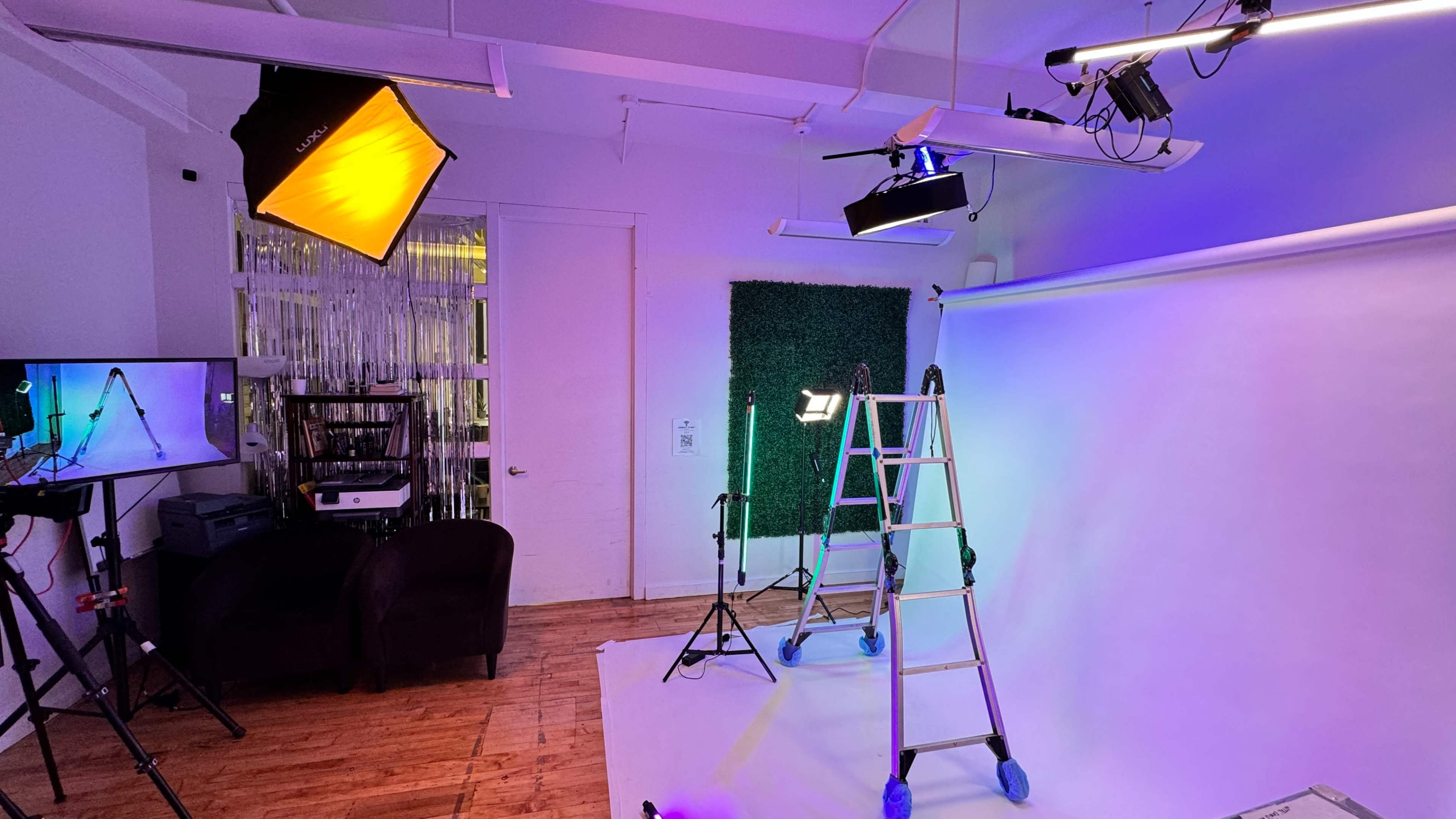 A photography studio setup features a ladder, multiple lighting fixtures, a backdrop, and a display monitor.