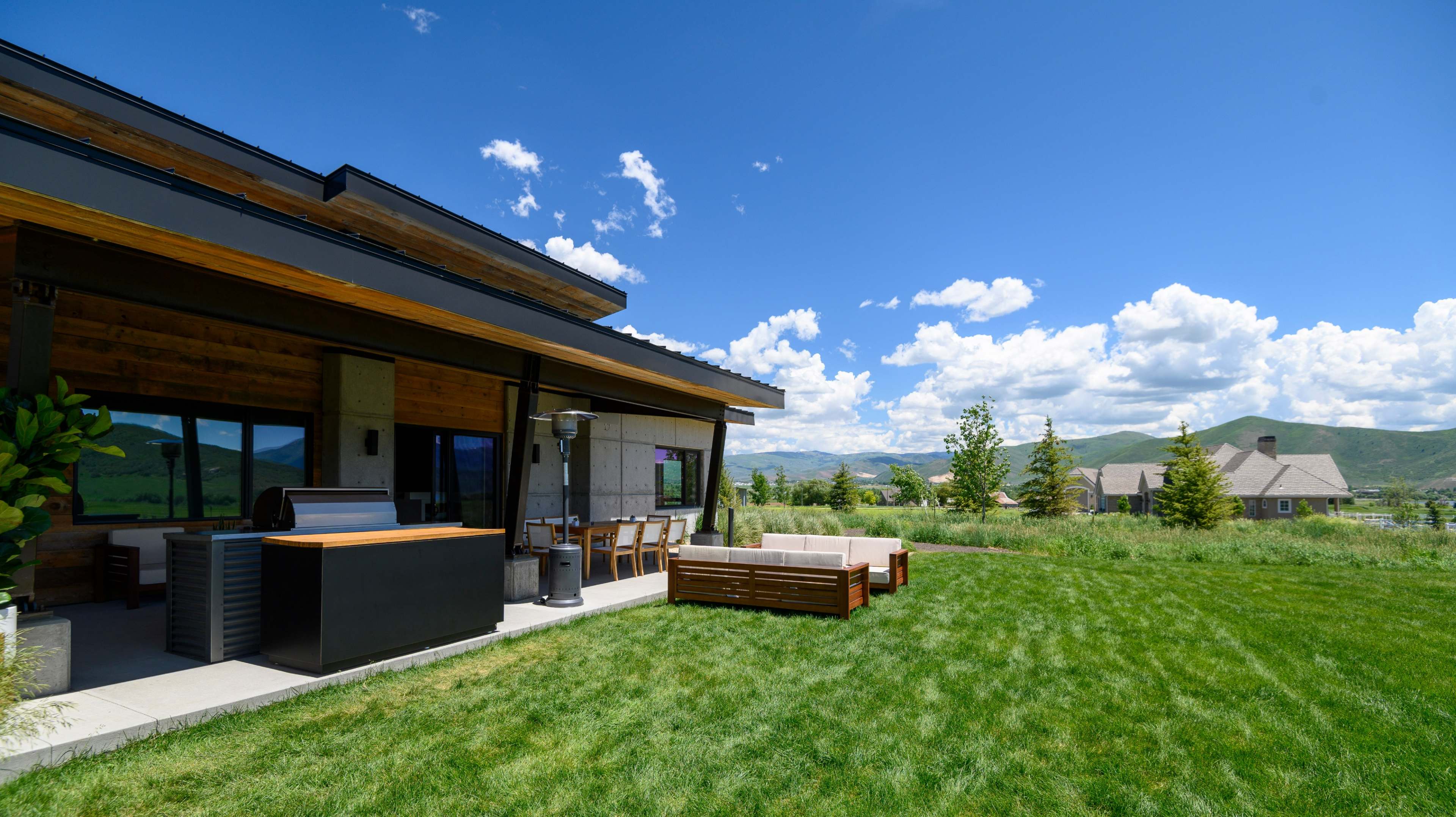 A modern house with a patio area featuring outdoor seating and a grill overlooks a green grassy landscape under a blue sky with clouds.