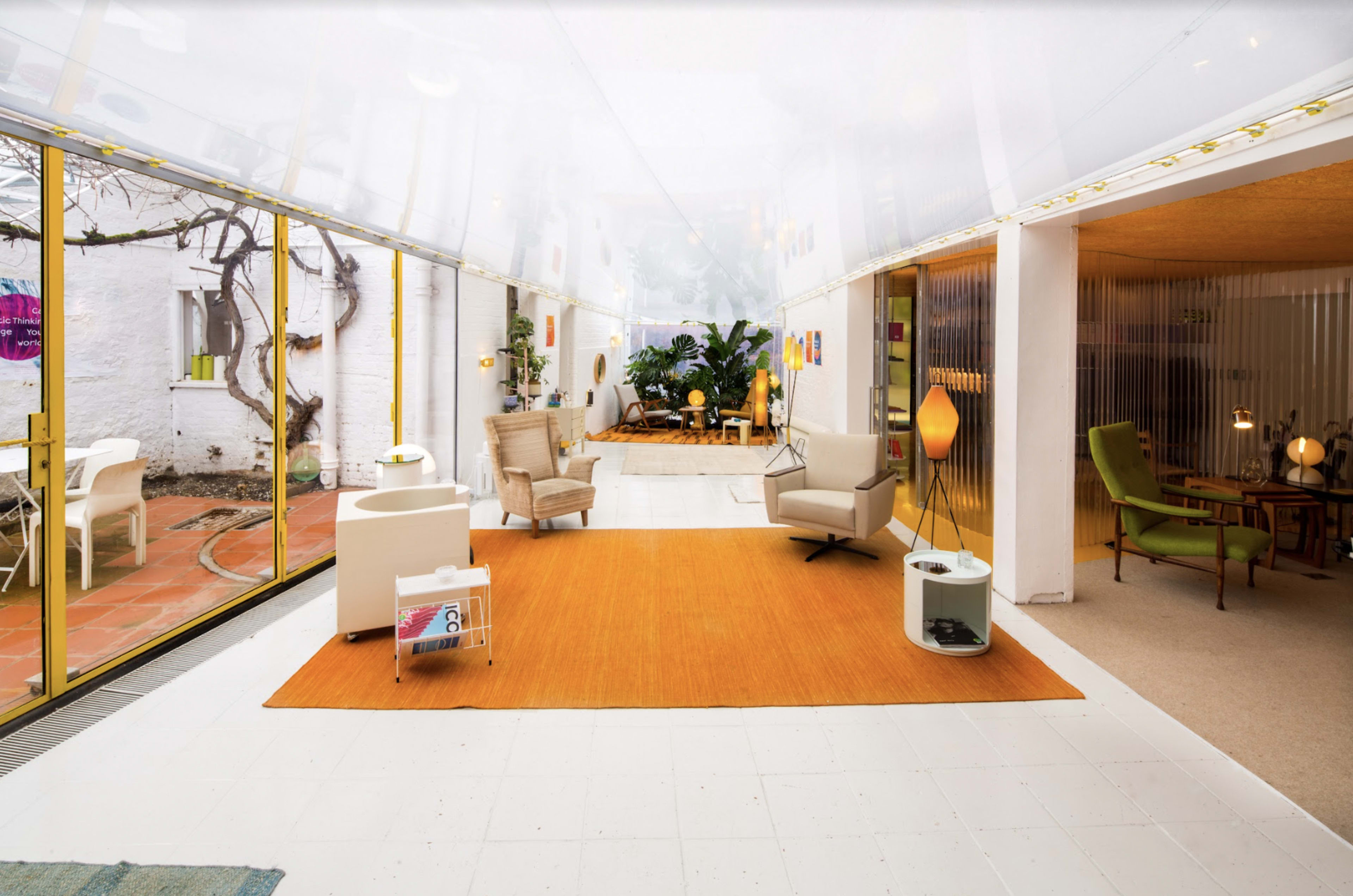 The image shows a spacious, modern interior with large glass walls, featuring a vibrant orange rug, various seating arrangements, and indoor plants.