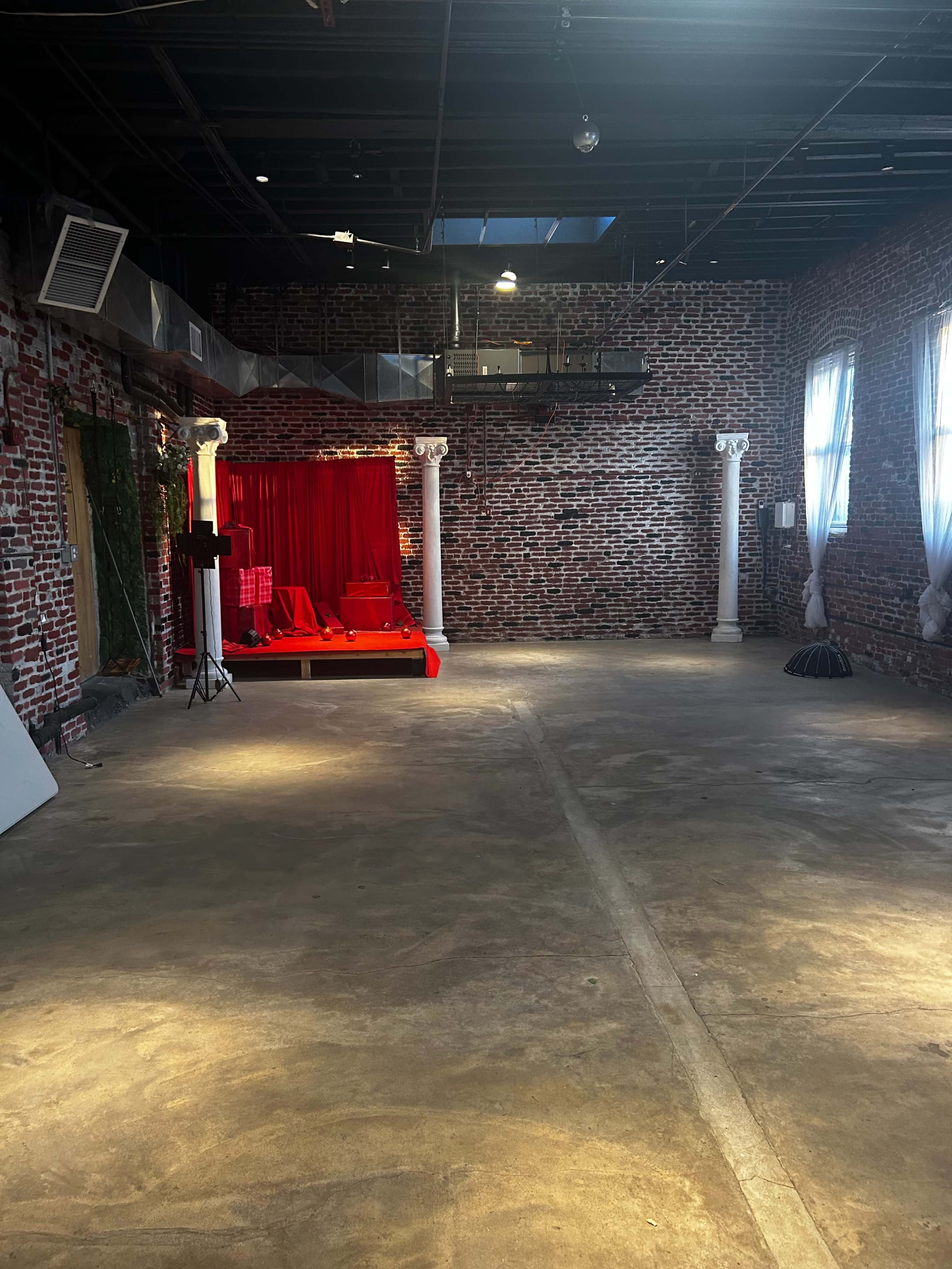 The image shows a spacious interior with exposed brick walls, a polished concrete floor, and a decorative red backdrop with drapery on one side.