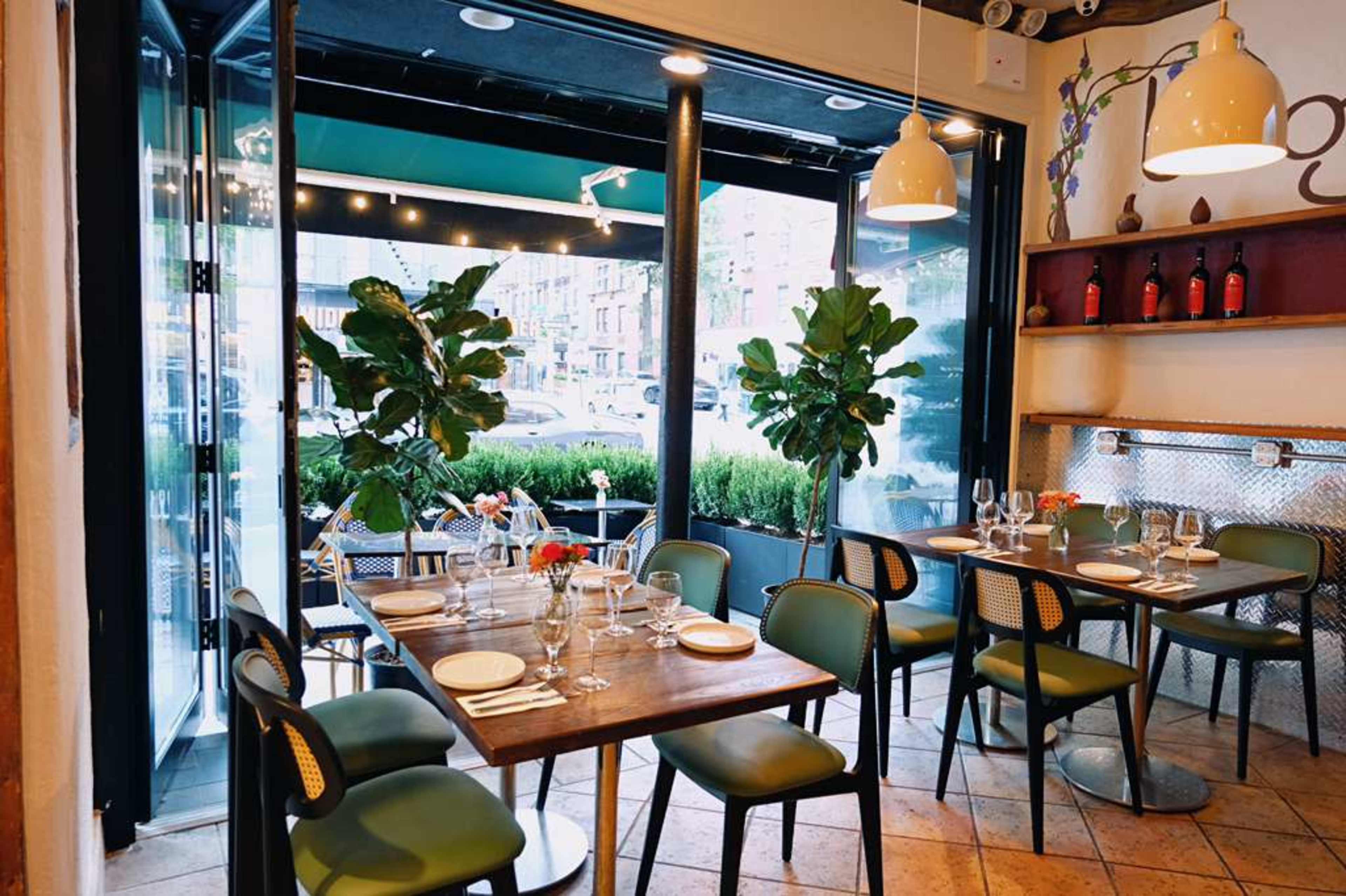 The image shows a stylish restaurant interior with tables set for dining, potted plants, and large windows displaying the street outside.