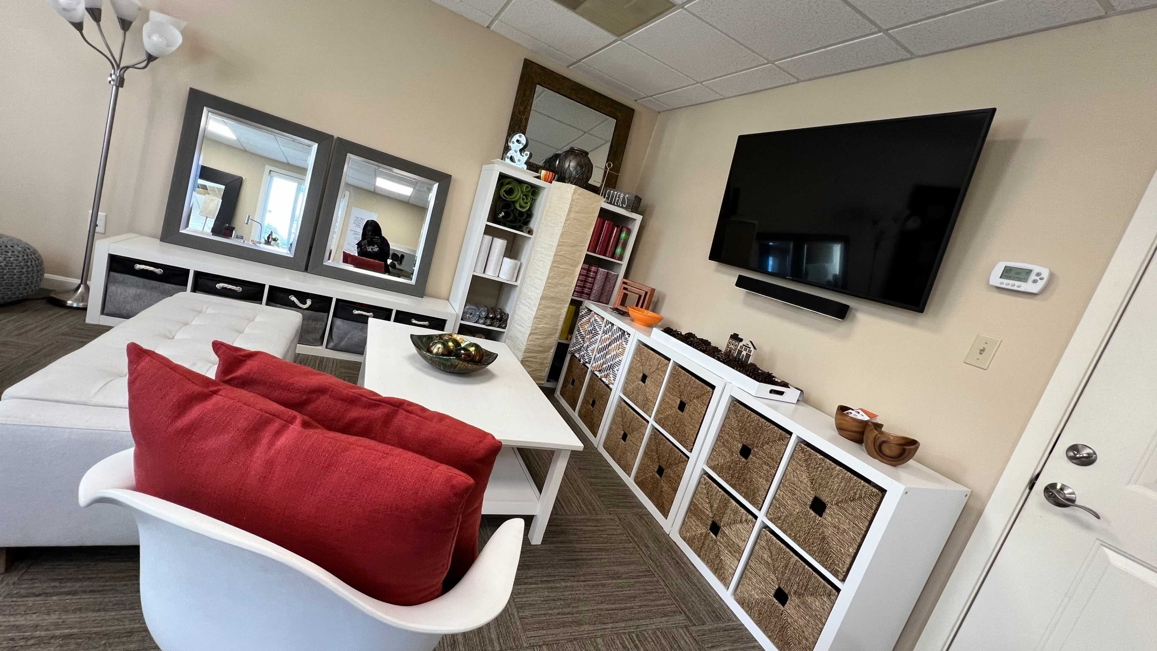 A modern office or lounge area with a white sofa, a white coffee table, storage cubbies, a wall-mounted television, and decorative items.