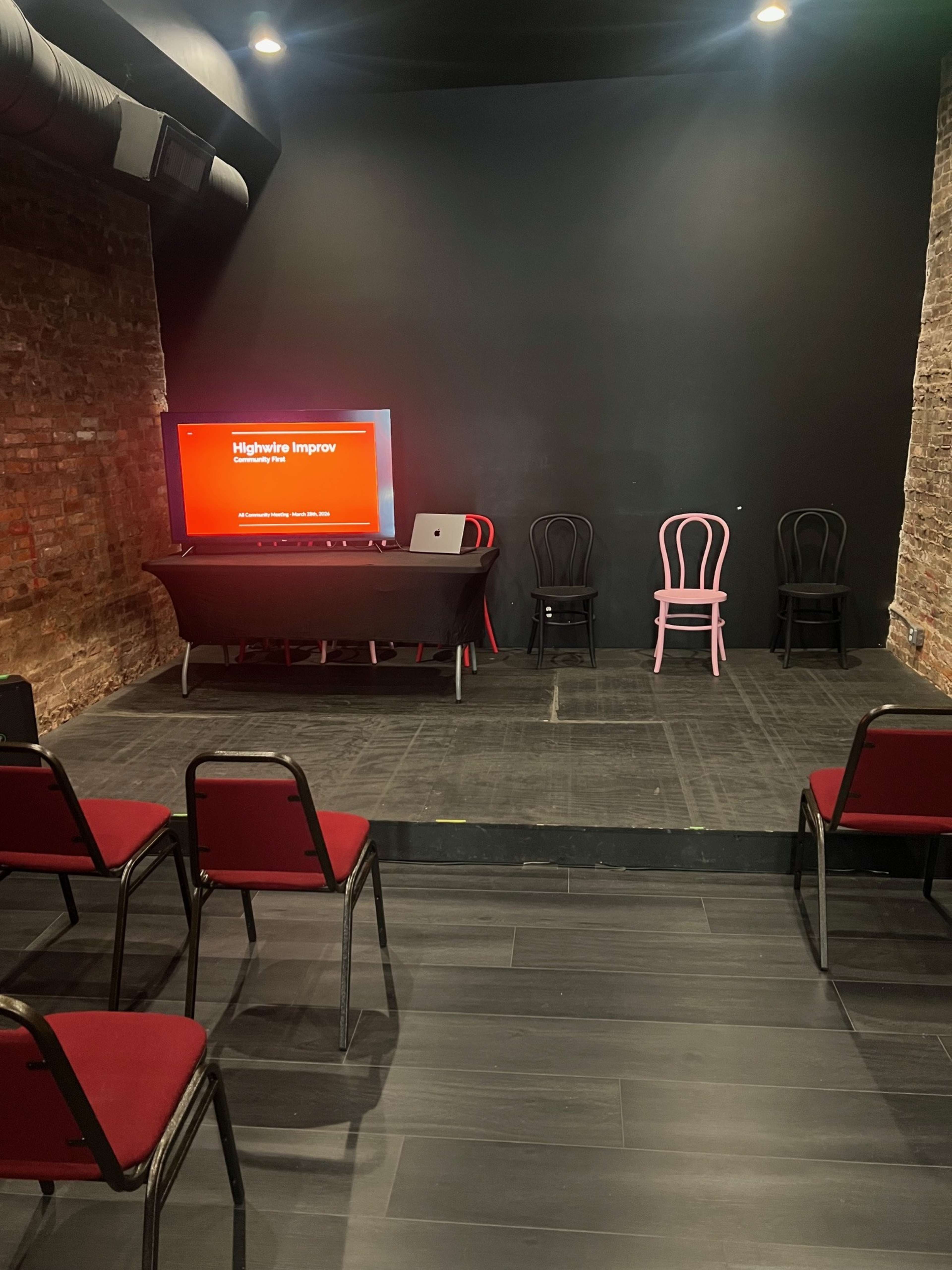 The image shows a small stage area with a black wall, a table holding a laptop and screen, and several chairs arranged in front of the stage.
