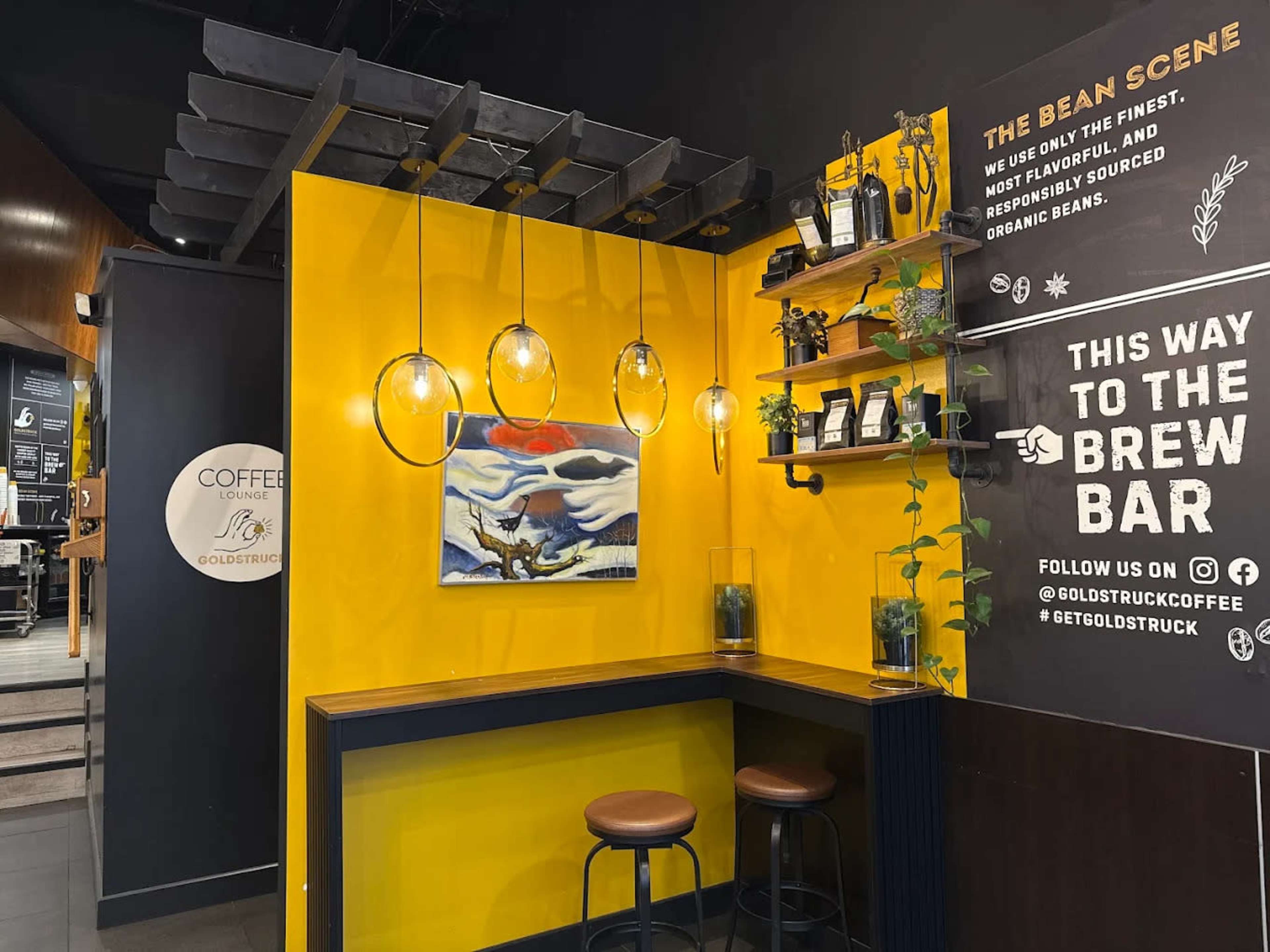 The image shows a cozy coffee shop corner with yellow walls, a wooden counter, pendant lights, and shelves filled with coffee-related items.
