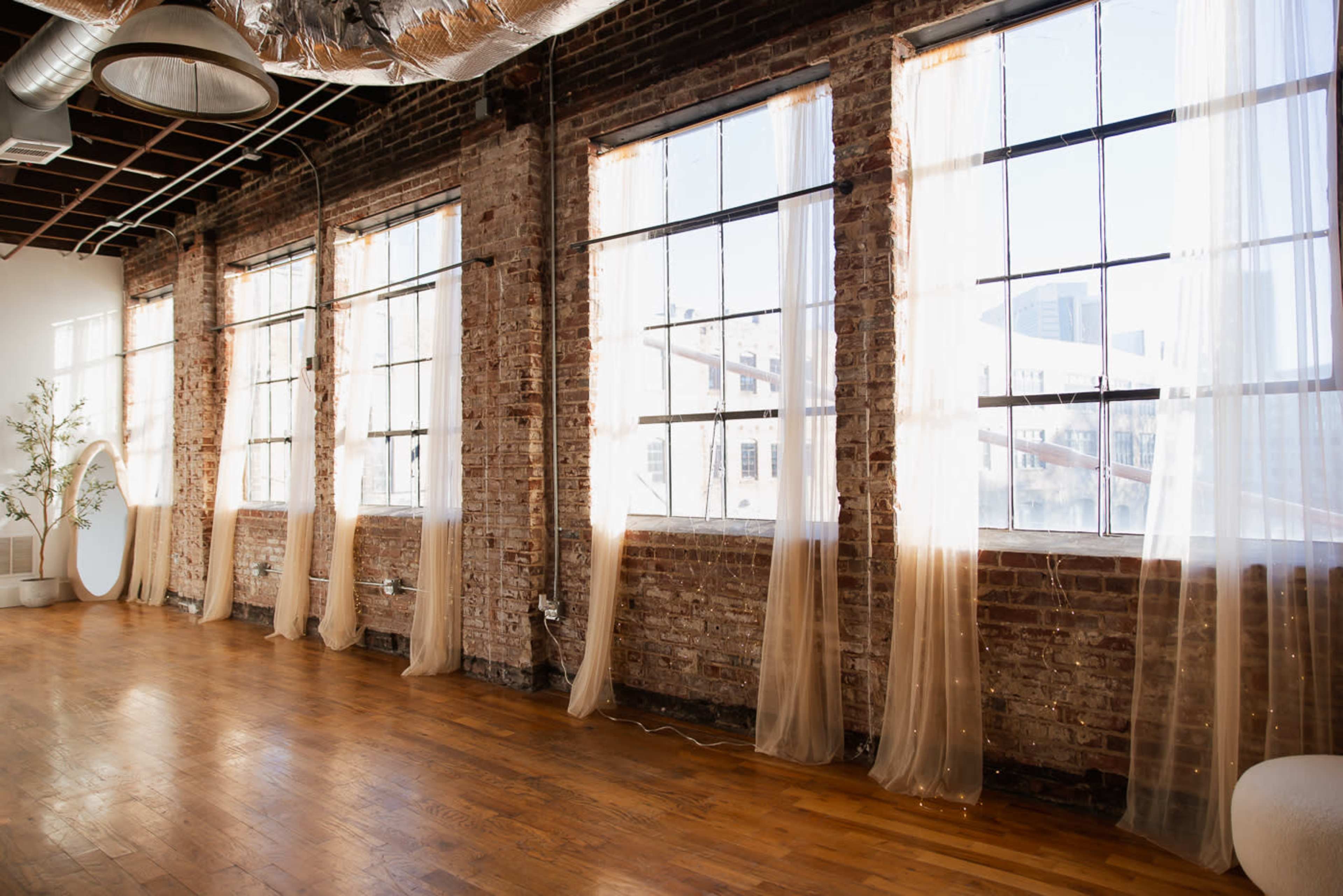 The image shows a spacious room with exposed brick walls, large windows draped with sheer curtains, and polished wooden flooring.