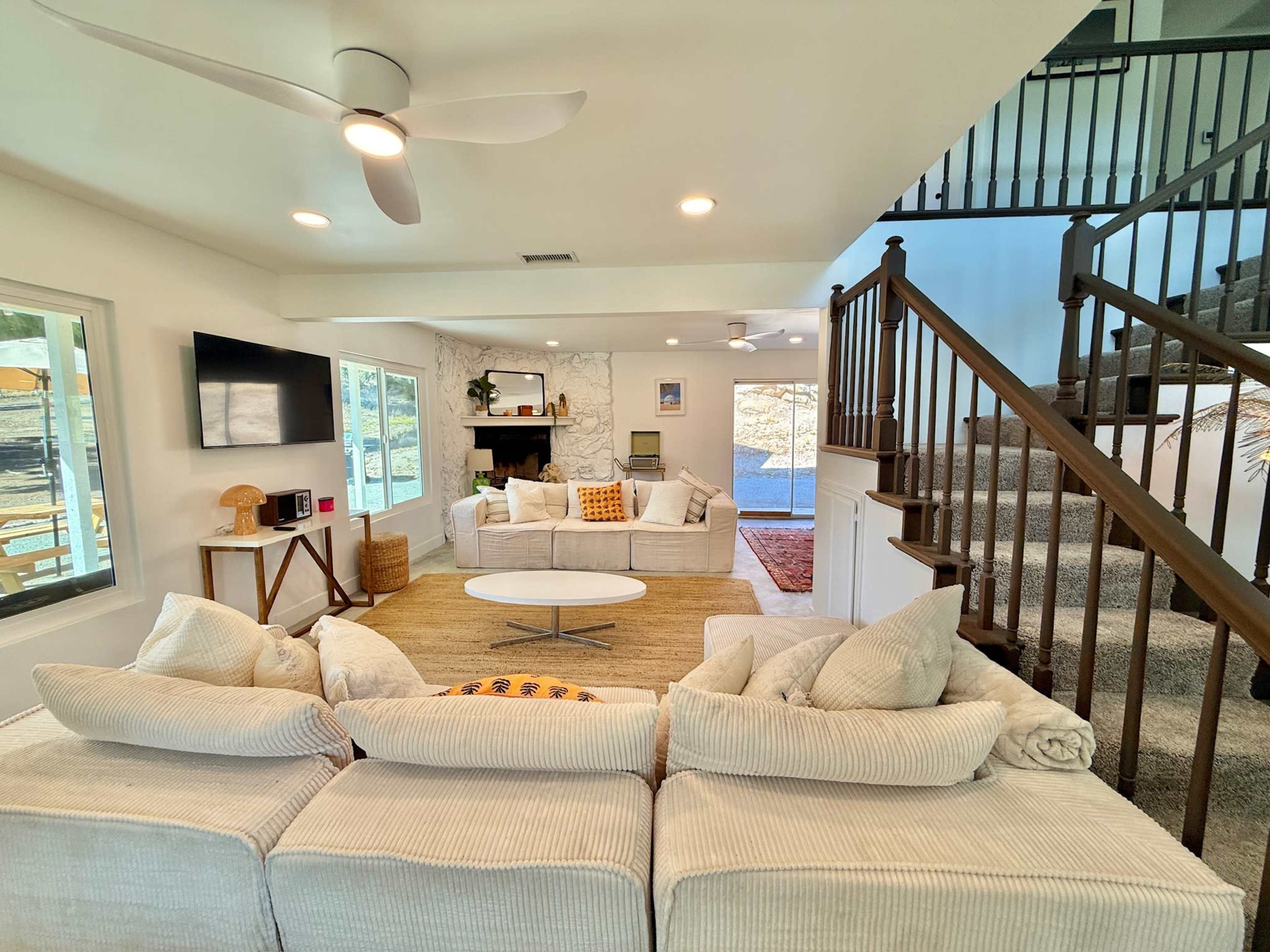 A bright living room features a white sectional sofa, a round coffee table, a fireplace, and a staircase leading to the upper level.