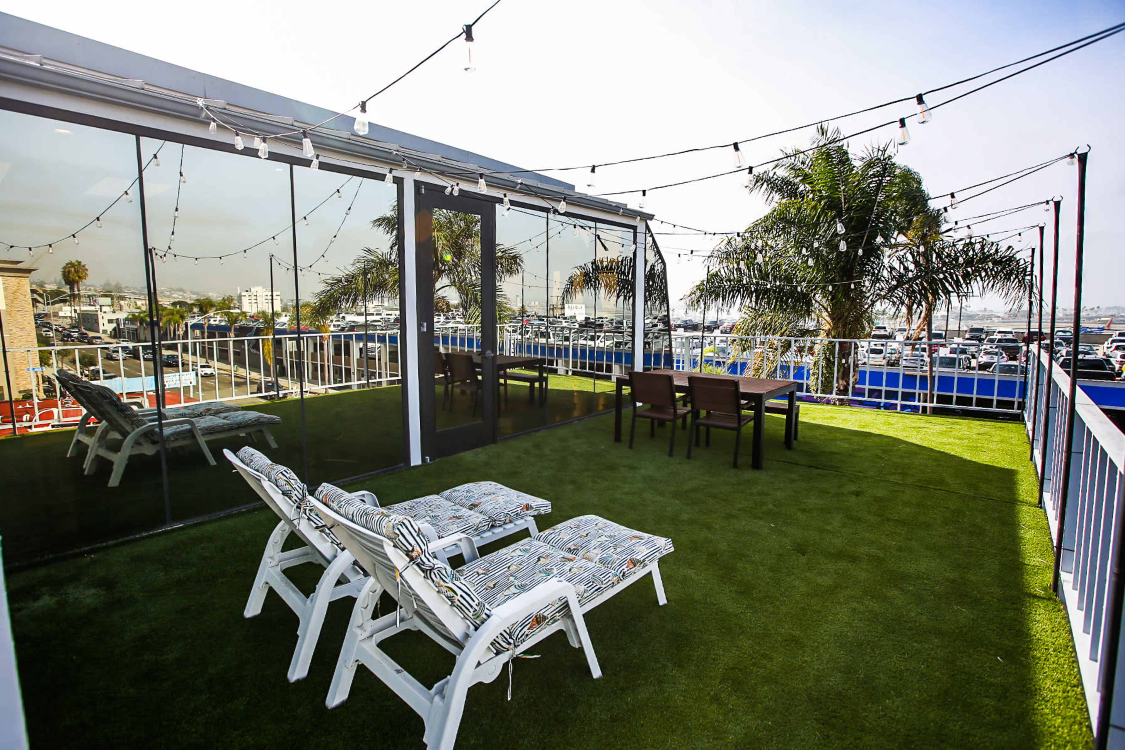 The image shows a rooftop space featuring two white lounge chairs, a glass-walled area, and a dining table surrounded by palm trees and string lights.