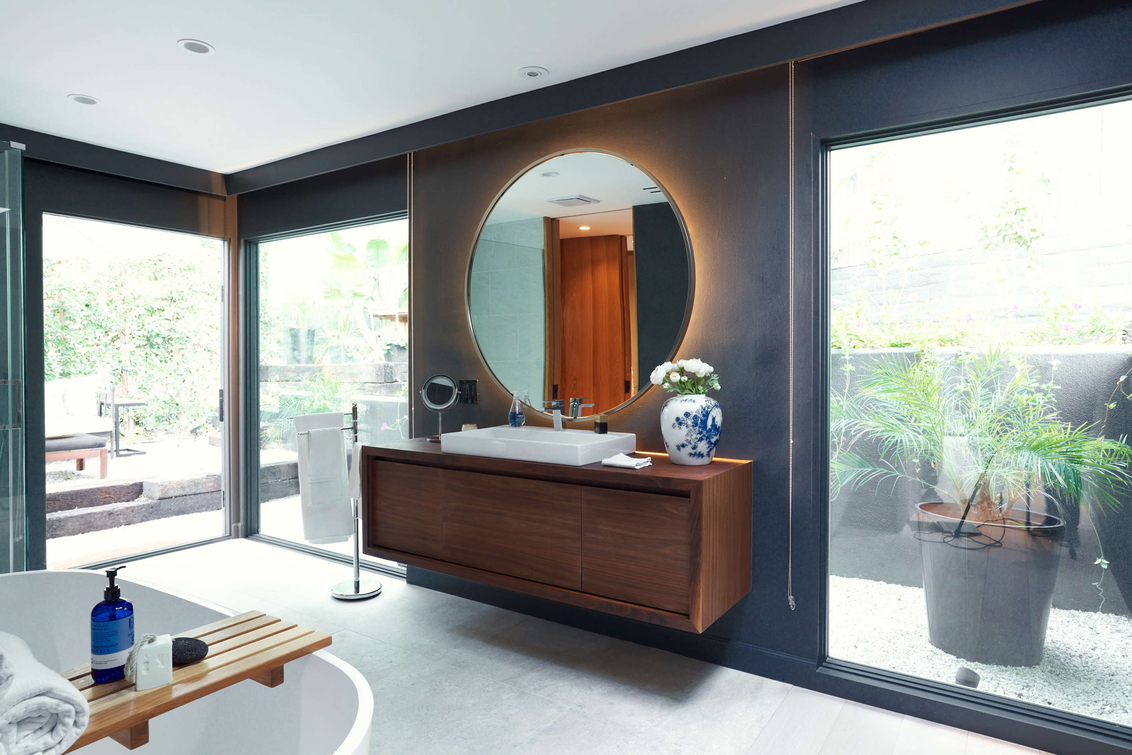 A modern bathroom features a wooden vanity with a round mirror, large windows providing views of an outdoor garden, and a freestanding bathtub.