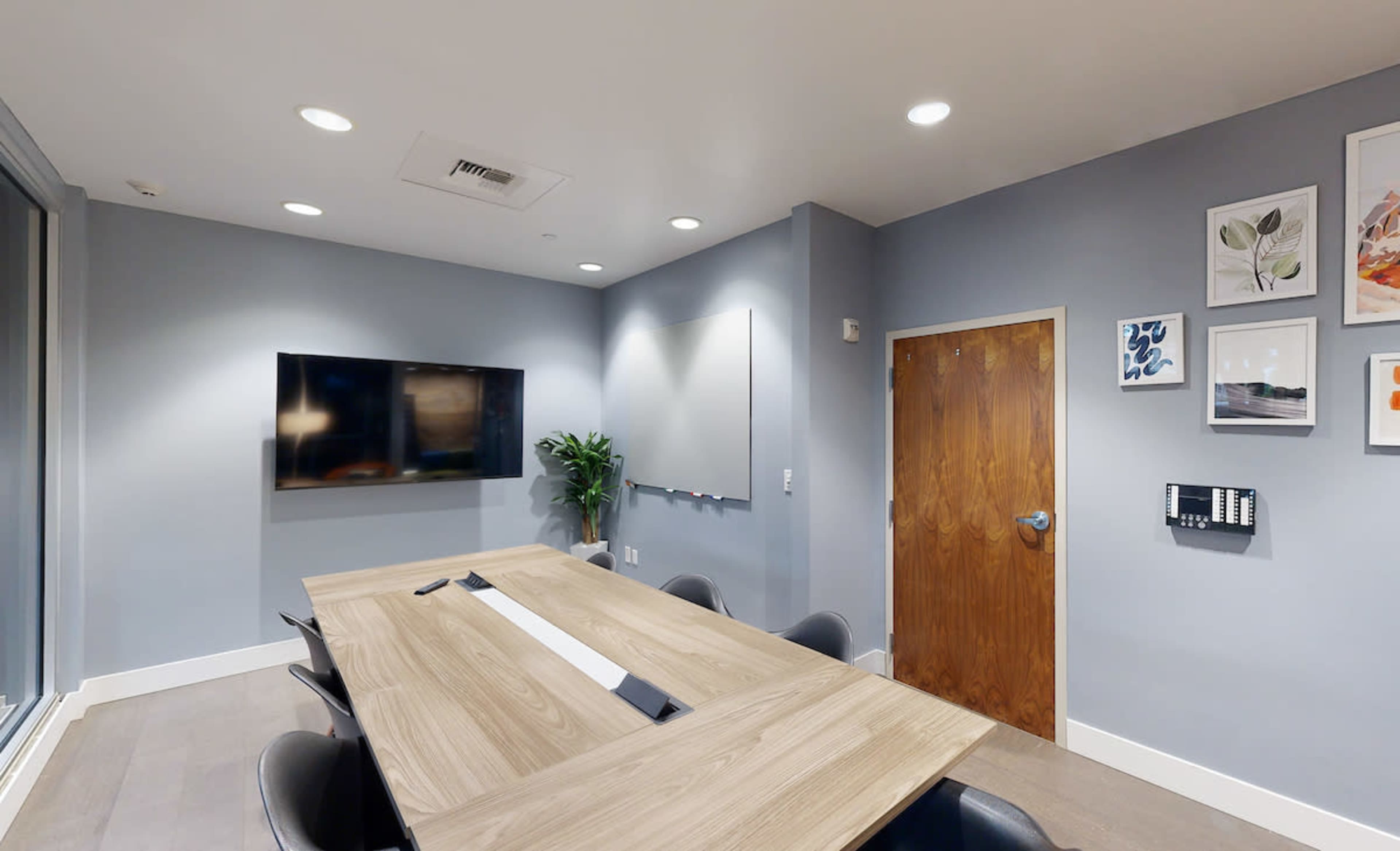 A modern conference room with a wooden table, black chairs, a wall-mounted television, and framed artwork on the walls.