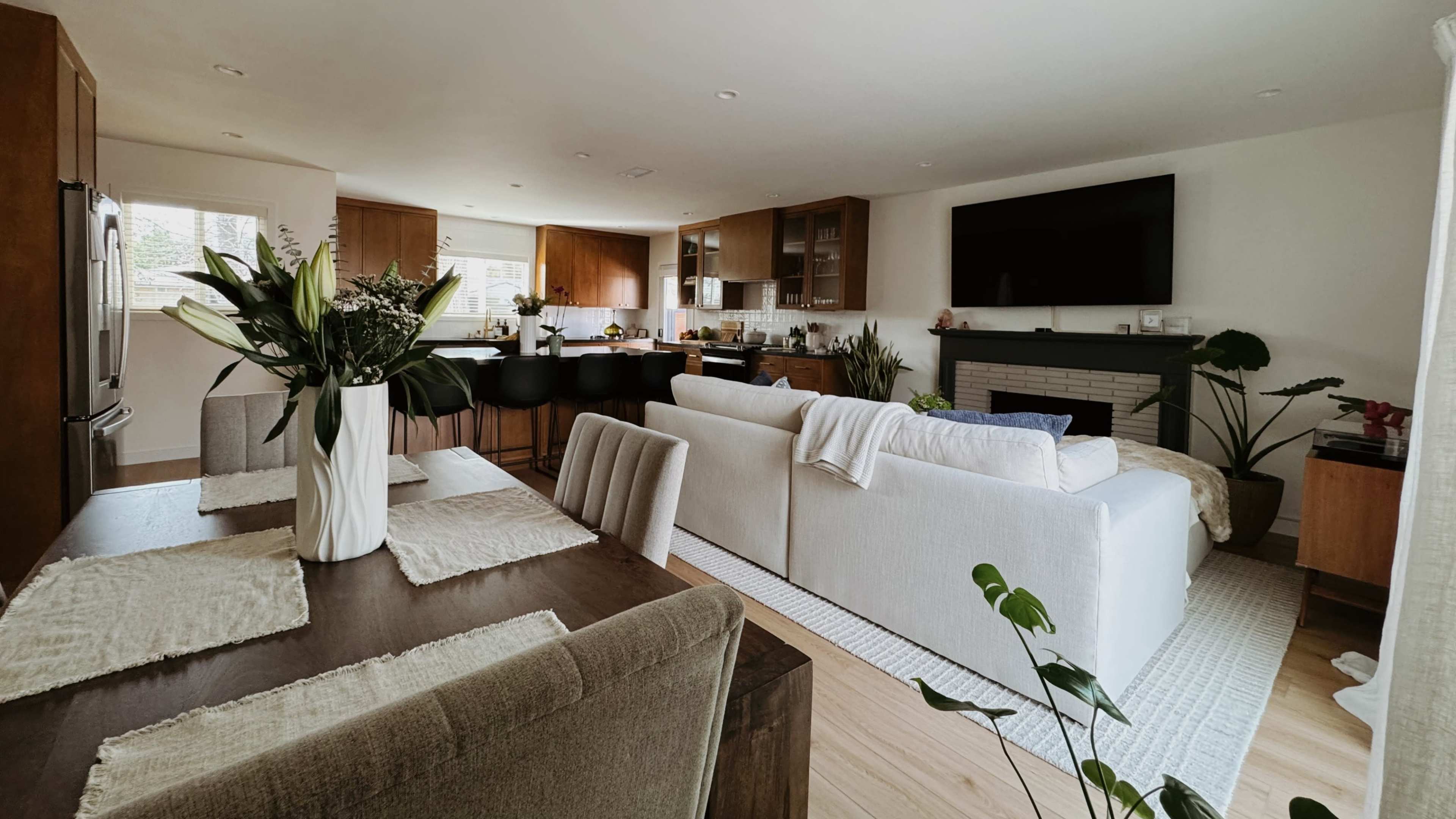 The image shows a modern interior of a living space featuring a dining table, a sofa, a kitchen area with bar seating, and large windows allowing natural light.