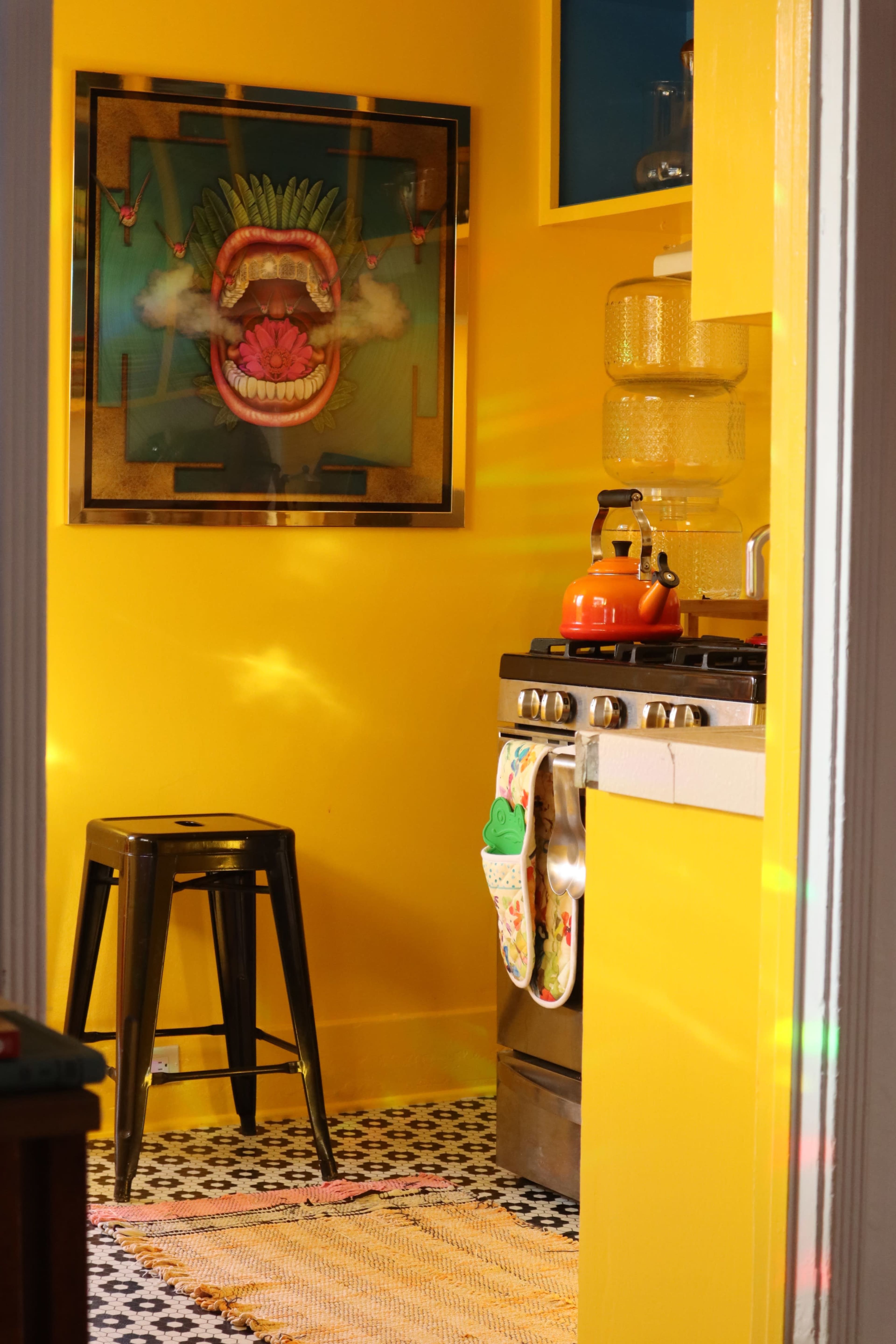 A brightly lit kitchen with yellow walls, featuring a stove, a black stool, and a colorful artwork of a mouth on the wall.