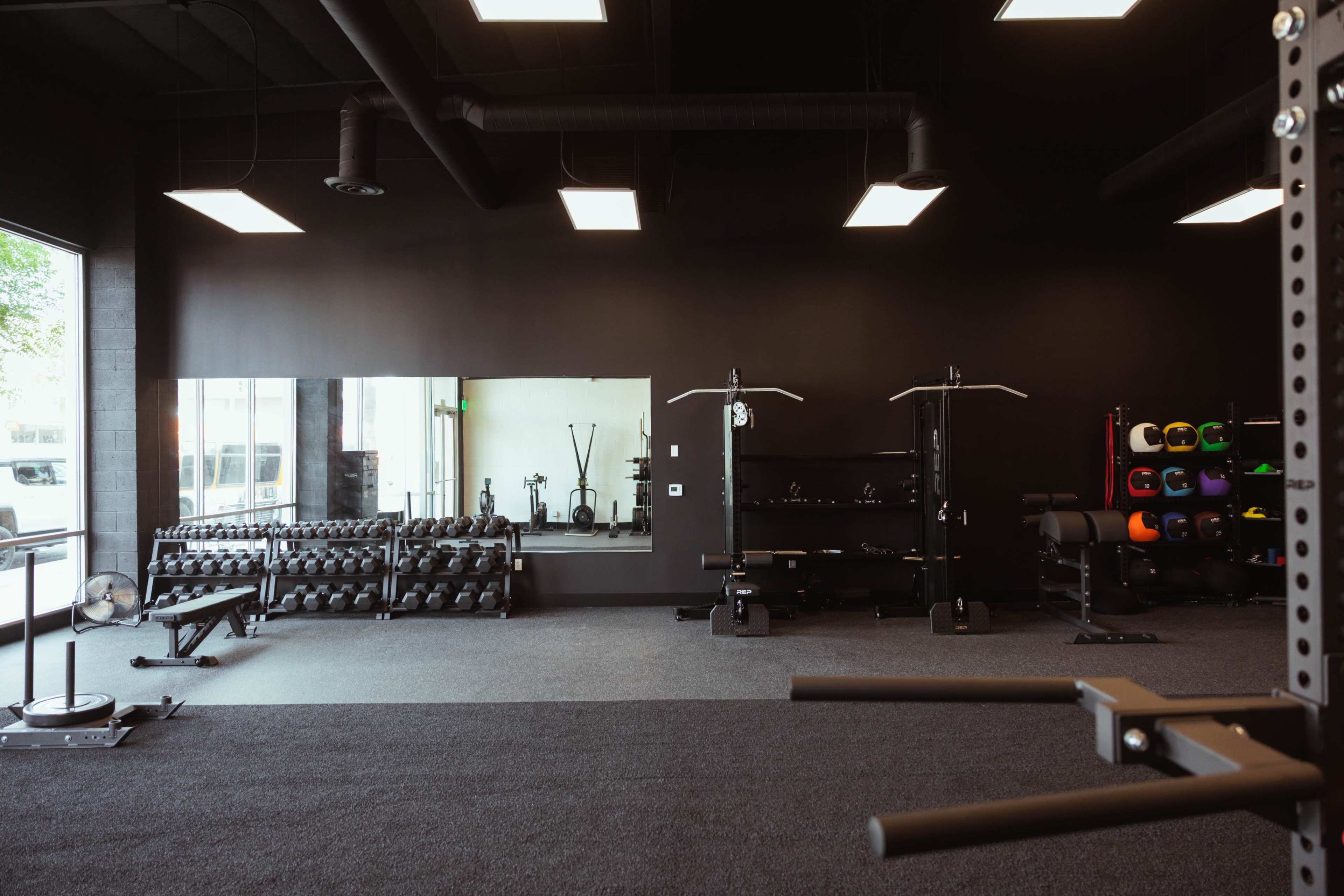 The image shows an empty gym with black walls, various weightlifting equipment, and a large mirror reflecting the space.