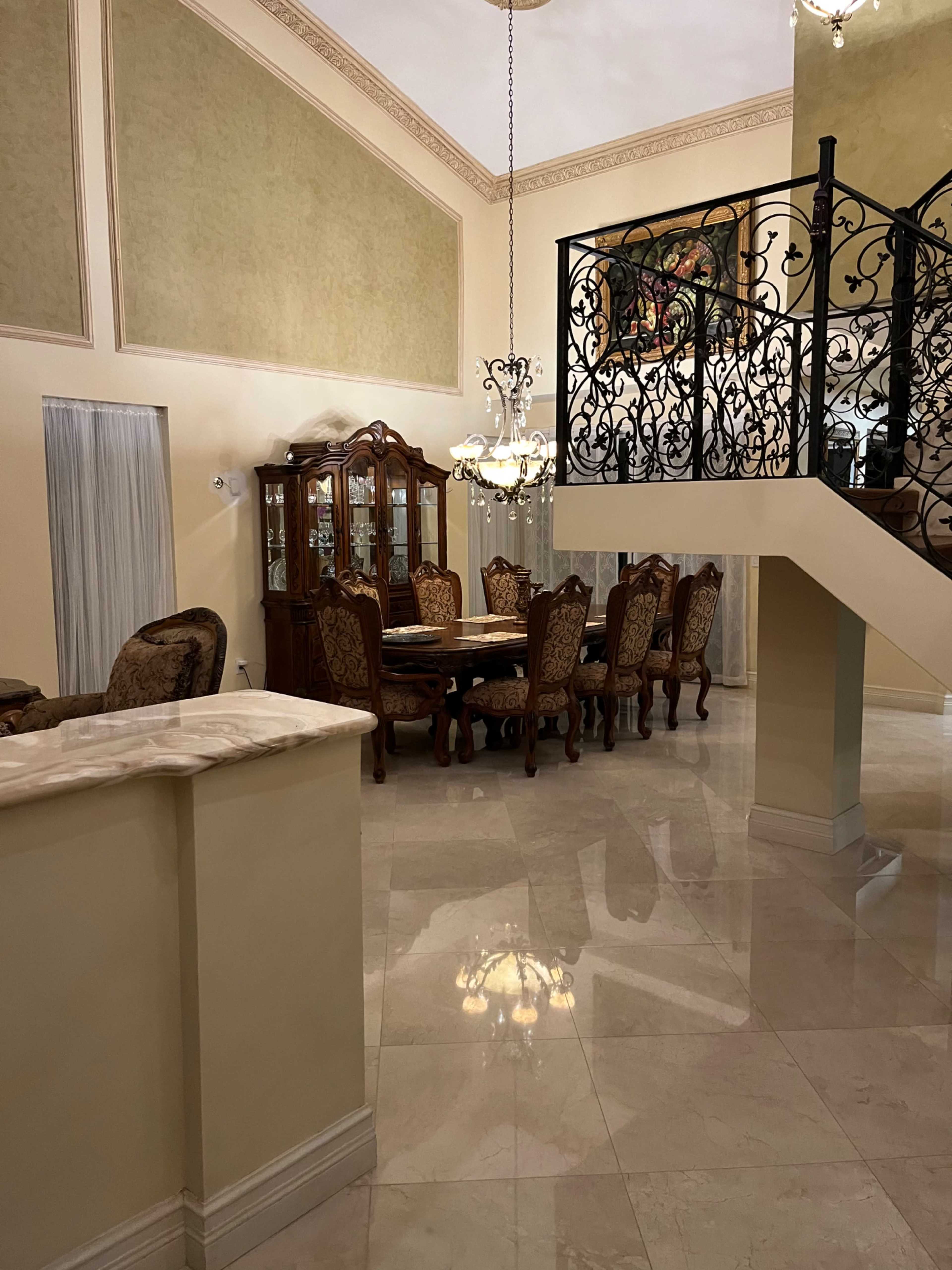 A spacious dining area with a large wooden table surrounded by ornate chairs, a glass-fronted china cabinet, and a staircase with decorative railing in a well-lit room.
